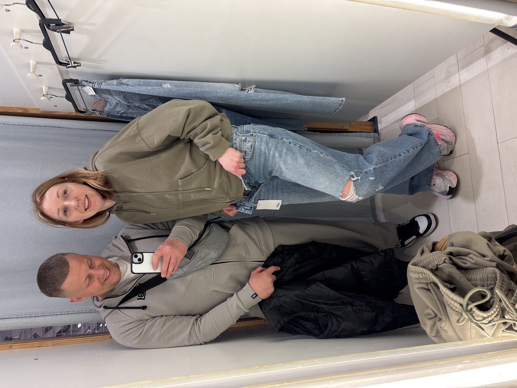 Two people are in a dressing room, a woman standing while trying on jeans and a man sitting on a bench taking a photo of them. The woman has medium-length blonde hair and is wearing a light green hoodie and distressed light wash jeans with pink and gray sneakers. The man has short brown hair and is wearing a light gray zip-up sweatshirt, a black crossbody bag, black and white sneakers, and is holding a white cellphone in his right hand. In the background, there are a pair of jeans on a hanger and folded on a shelf, as well as other articles of clothing on the bench near the man. The dressing room has light gray walls and light-colored wood trim. There is no indication of a city or country in the image.