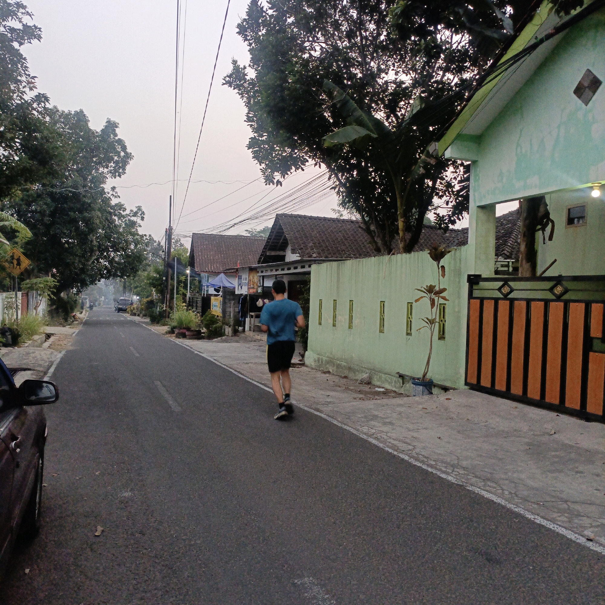 A man is jogging on a paved road in an urban area. He is wearing a blue t-shirt and black shorts. The road is lined with houses and trees, and power lines are visible overhead. A car is parked on the side of the road. The sky is overcast. The scene appears to be in Salatiga, Indonesia.