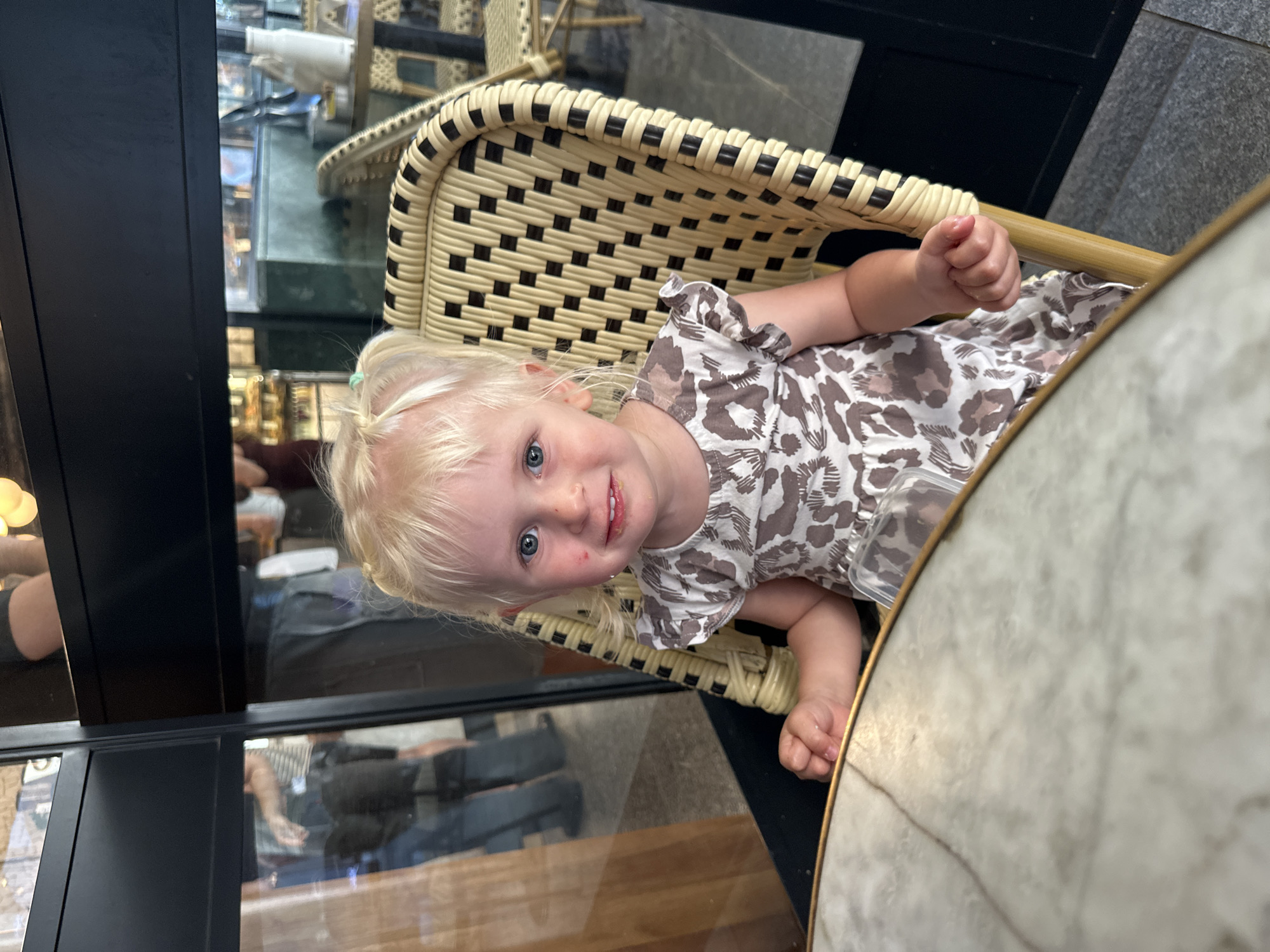 A young girl with blonde hair tied in pigtails and blue eyes is seated at a table in what appears to be a cafe or restaurant. She is wearing a short-sleeved dress with a leopard-like print in shades of brown and white. Her right hand is resting on the table, and her left hand is on her lap. The table has a marble top with gold trim. Behind her, a rattan chair with a woven pattern is visible. In the background, through what looks like glass partitions, other people and cafe furniture can be seen. The setting suggests an indoor area with outdoor seating visible beyond. The overall scene depicts a casual dining environment. The city of Herzliya, Israel, is a coastal city known for its beaches, upscale residential areas, and technology hubs, and it features many cafes and restaurants that could match this environment.