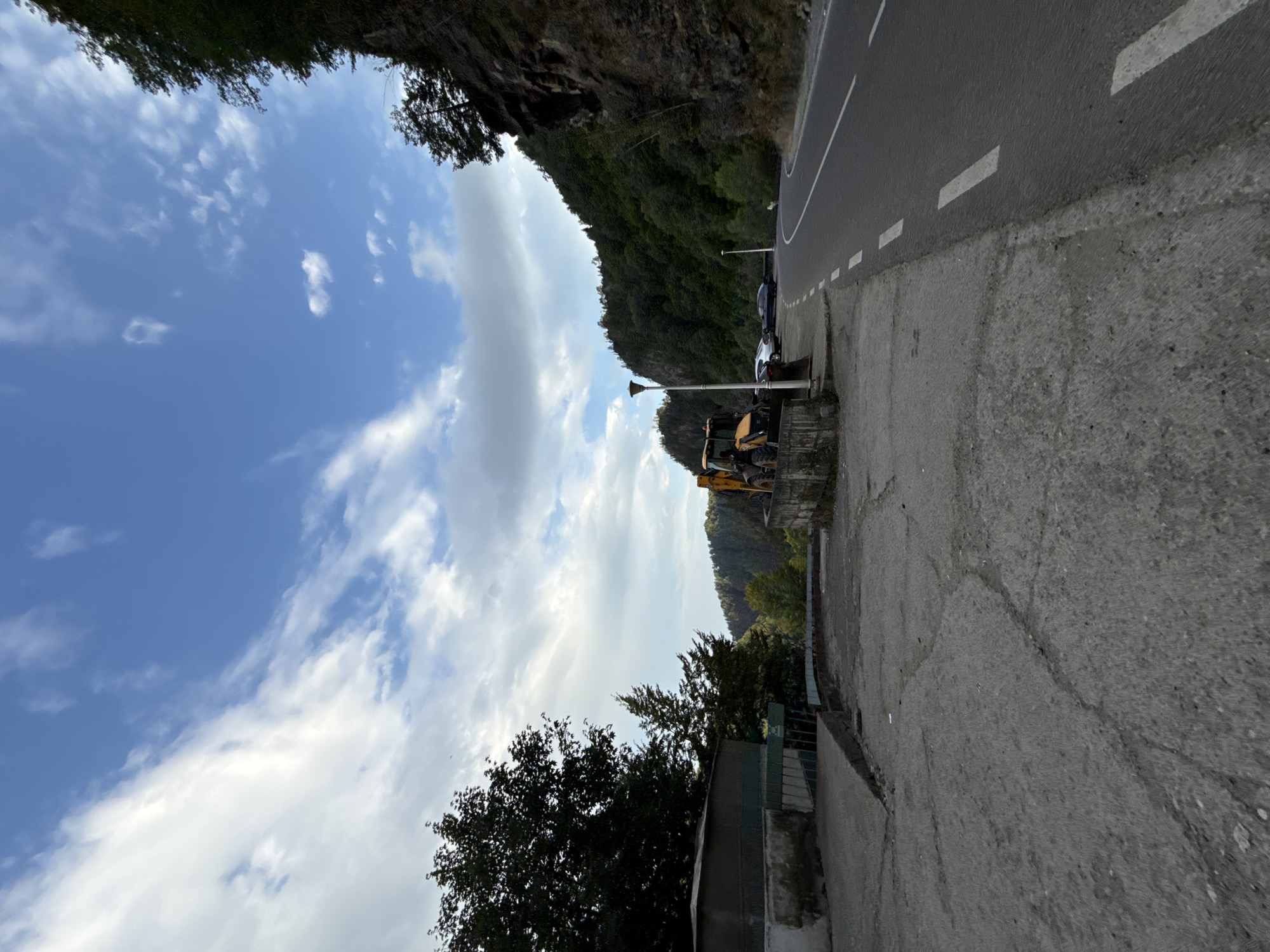 The image depicts a scenic view on a cloudy day, likely in Arefu, Romania, given the mountainous terrain and road infrastructure suggestive of such a location. In the foreground, a cracked concrete surface leads towards a paved road that curves into the distance. To the left, a low concrete wall runs alongside the road, bordering a gravelly area with a green structure that appears to be a small building or shed. A yellow backhoe loader is parked on this gravelly area, next to a concrete pedestal. Further back, several cars are parked on the side of the road. The background is dominated by a steep, tree-covered mountainside on the right and a dense forest on the left, with a vast blue sky filled with wispy clouds above. The overall impression is one of a tranquil, natural setting with evidence of human activity, possibly related to construction or maintenance work given the presence of the heavy machinery.