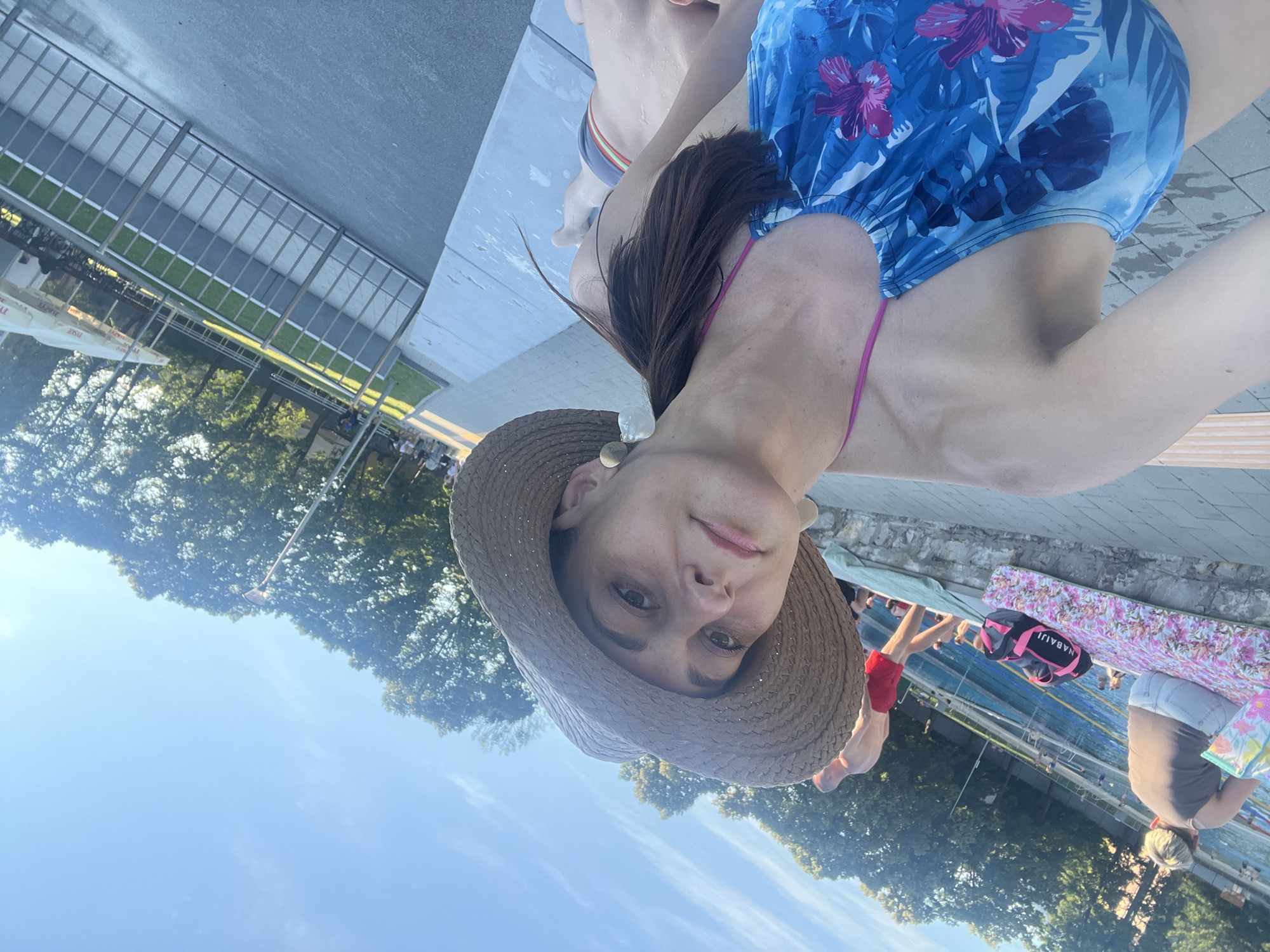 A woman wearing a blue floral print swimsuit and a straw hat is taking a selfie at an outdoor swimming pool. She is sitting on a gray concrete ledge with a body of water in the background. There are several other people in the background, some in the pool and some on the side. The weather appears to be sunny.
