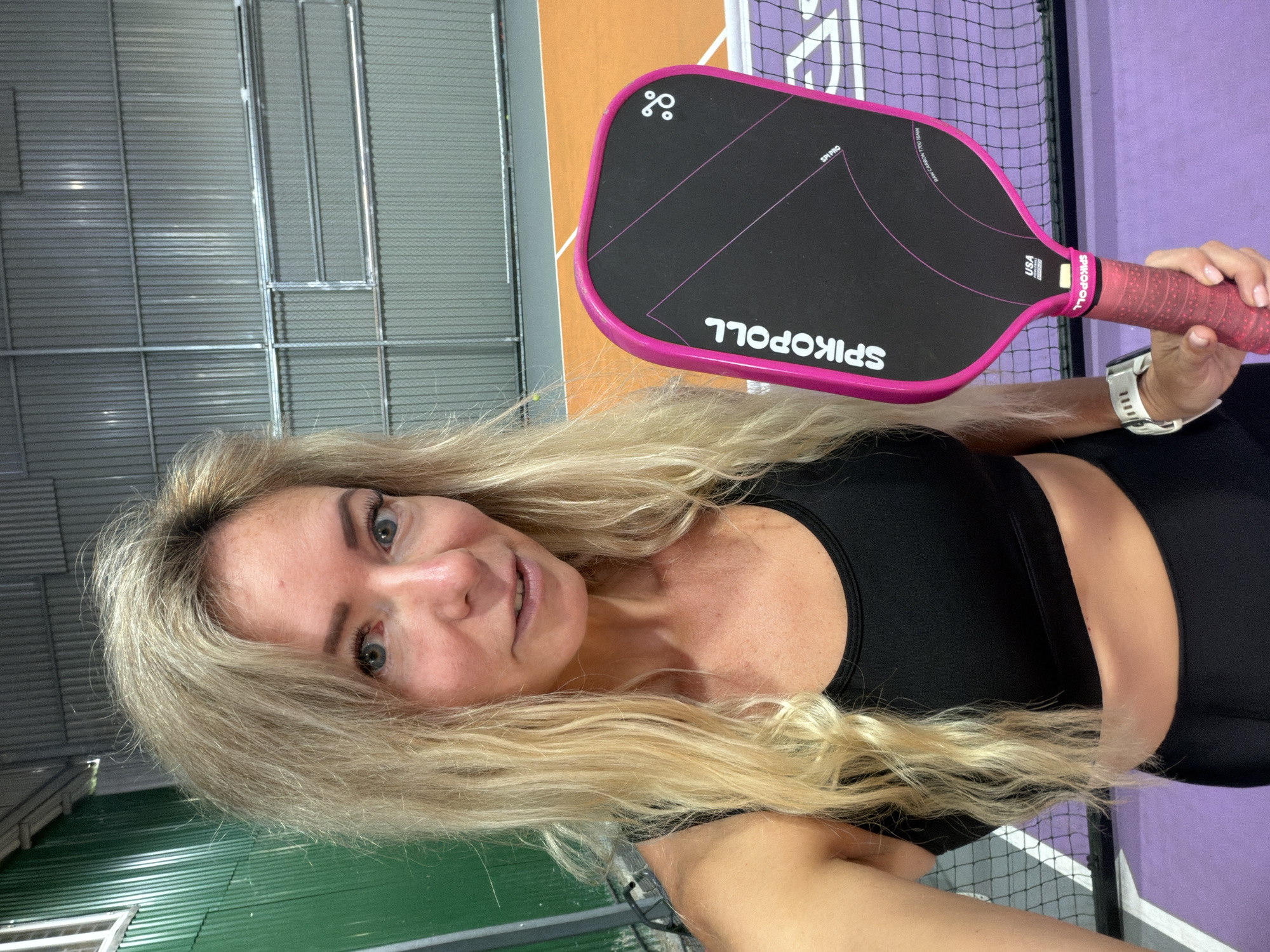 In an indoor pickleball court, a woman with long, blonde, wavy hair is taking a selfie. She is wearing a black sports bra and black athletic shorts, with a white smartwatch on her left wrist. She is holding a pink and black pickleball paddle with the brand name "SPIKOPOLL" visible. The court has purple and orange flooring, with a net in the foreground and a textured metal wall in the background. The location is an indoor sports facility. There is no indication that the location is in Da Lat, Vietnam.