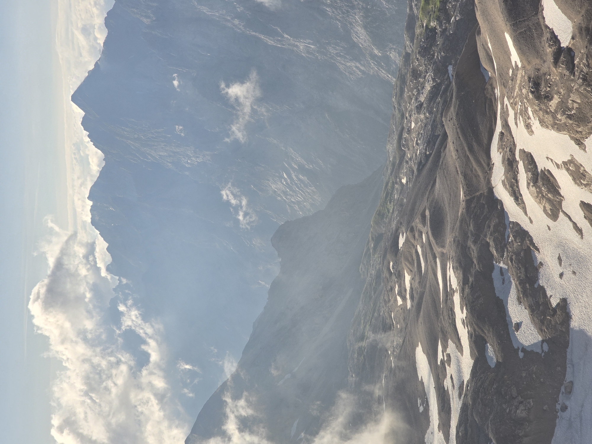 The image depicts a panoramic view of the Bavarian Alps near Garmisch-Partenkirchen, Germany. The foreground is a rocky, uneven terrain with patches of snow scattered across it. In the middle ground, rolling hills and slopes rise, also interspersed with snow. The background is dominated by majestic, jagged mountain peaks that fade into a hazy blue due to atmospheric perspective. Wispy clouds drift amongst the mountains and in the sky above, adding a sense of depth and vastness to the landscape. The lighting suggests it is either early morning or late afternoon, with soft sunlight illuminating the scene. There are no people or man-made objects visible, focusing solely on the natural grandeur of the alpine environment.