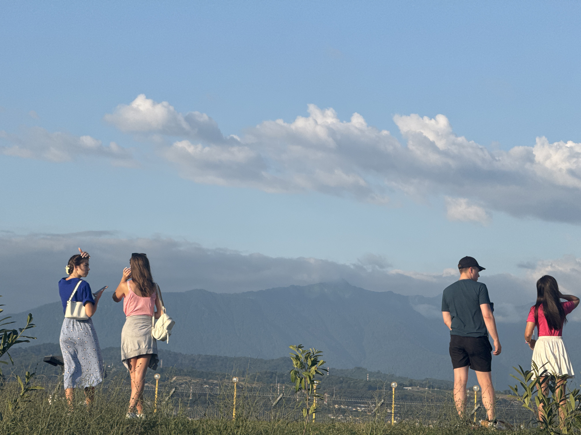 Four people stand on a grassy hill, facing away from the camera towards a mountain range under a bright blue sky with scattered white clouds. The sun appears to be setting or rising, casting a warm light on the scene. The person on the far left wears a blue short-sleeved shirt and a blue patterned skirt, carrying a white purse. Next to them, a person in a pink tank top and gray skirt holds a white purse. To the right, a man in a dark blue t-shirt and black shorts wears a black baseball cap. The person on the far right is a woman in a bright pink t-shirt and a white skirt. The background shows rolling hills leading to a majestic mountain range, with some distant structures and lights visible in the valley below. The overall mood is relaxed and contemplative, with the individuals seemingly enjoying the scenic view. No visible text is present in the image.