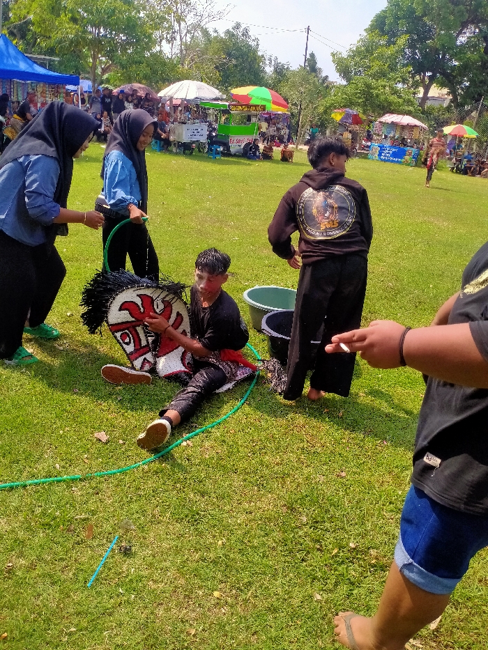 This image captures an outdoor event in Jetak, Indonesia, on a sunny day. In the foreground, five individuals are situated on a bright green grassy field. A male subject is seated on the grass, facing slightly right, wearing a wet black t-shirt, black pants, and brown and white shoes. He has red markings on his face and is holding a large, white and red stylized mask with a black, spiky mane. A red fabric sash is visible around his waist. Two females, both wearing hijabs (one in a blue long-sleeved shirt, the other in a light blue long-sleeved shirt), stand to his left, actively hosing him and the mask with water from a green garden hose. Behind the seated male, another male stands facing away from the camera, wearing a black hooded sweatshirt with a circular gold emblem on the back and black pants, barefoot. On the far right, partially visible, a person's arm and lower torso are shown; this individual holds a lit cigarette and wears a black t-shirt, blue denim shorts, and flip-flops. Two plastic buckets, one green and one black, are on the grass behind the seated male. A blue plastic straw lies on the grass in the lower-center.
The midground features several market stalls or vendor booths arranged across the field. These stalls are covered by various colorful umbrellas (white, red, multi-colored) and canopies (including a prominent blue one on the left). People are visible congregating around these stalls. One stall prominently displays "ES SOY." The background consists of dense green trees under a clear blue sky, with visible utility poles and power lines.