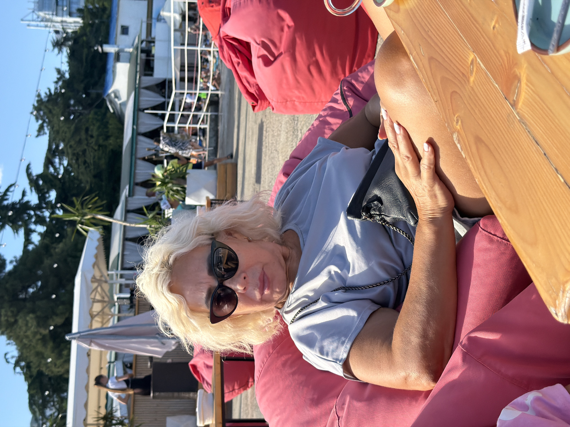 A medium shot shows a woman with blonde, curly hair sitting in a large, pink beanbag chair on a sunny day at a beachside establishment in Alushta, Ukraine. She is wearing large, black, cat-eye sunglasses and a light grey t-shirt. A black clutch purse hangs from her shoulder, and her right hand rests on her leg. She is positioned in front of a wooden table. In the background, there are more beanbag chairs, beach umbrellas, and people sitting at tables. Trees and buildings are visible further in the distance, with a clear blue sky overhead.