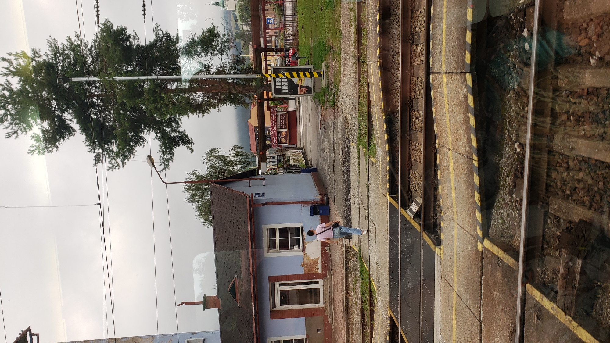 The image depicts a train station in Likavka, Slovakia. A woman wearing a pink t-shirt and blue jeans is walking away from a light blue building with a brown roof. She is carrying a black bag over her shoulder. In the foreground, there are railroad tracks with yellow and black striped crossing barriers. In the background, there are more buildings, trees, and a distant church steeple. A sign with "HORKO DOKONCE" is visible on a pole. The sky is overcast.