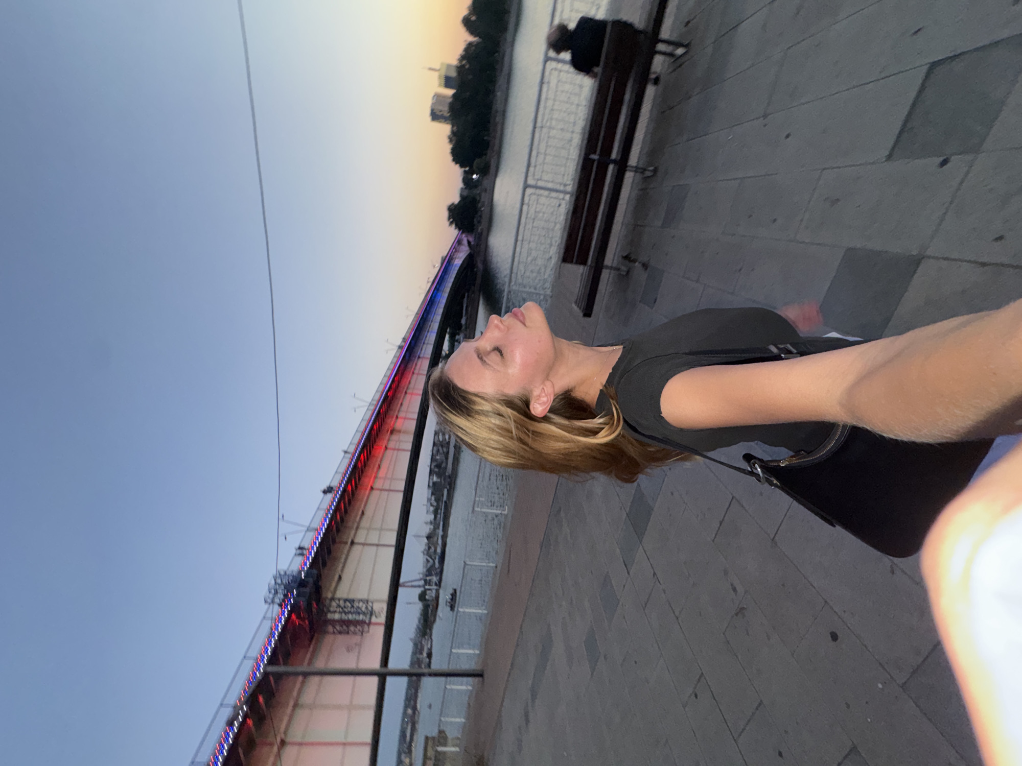 In an outdoor, low-light shot captured at dusk, a young woman with blonde hair is positioned in the foreground, facing right with her eyes closed and head tilted back, seemingly basking in the ambient light. She is wearing a dark t-shirt and carrying a black purse on her left shoulder. The background features a wide river with a brightly lit bridge spanning across it, displaying red and blue lights. The bridge's structure is a prominent element, with lights illuminating its railing and undersides. Along the riverbank, a paved walkway is visible, with a park bench occupied by a person in silhouette. In the distance, city buildings and trees are silhouetted against the fading sky. The overall scene suggests a public area along the Sava River in Belgrade, Serbia, near the Branko's Bridge.