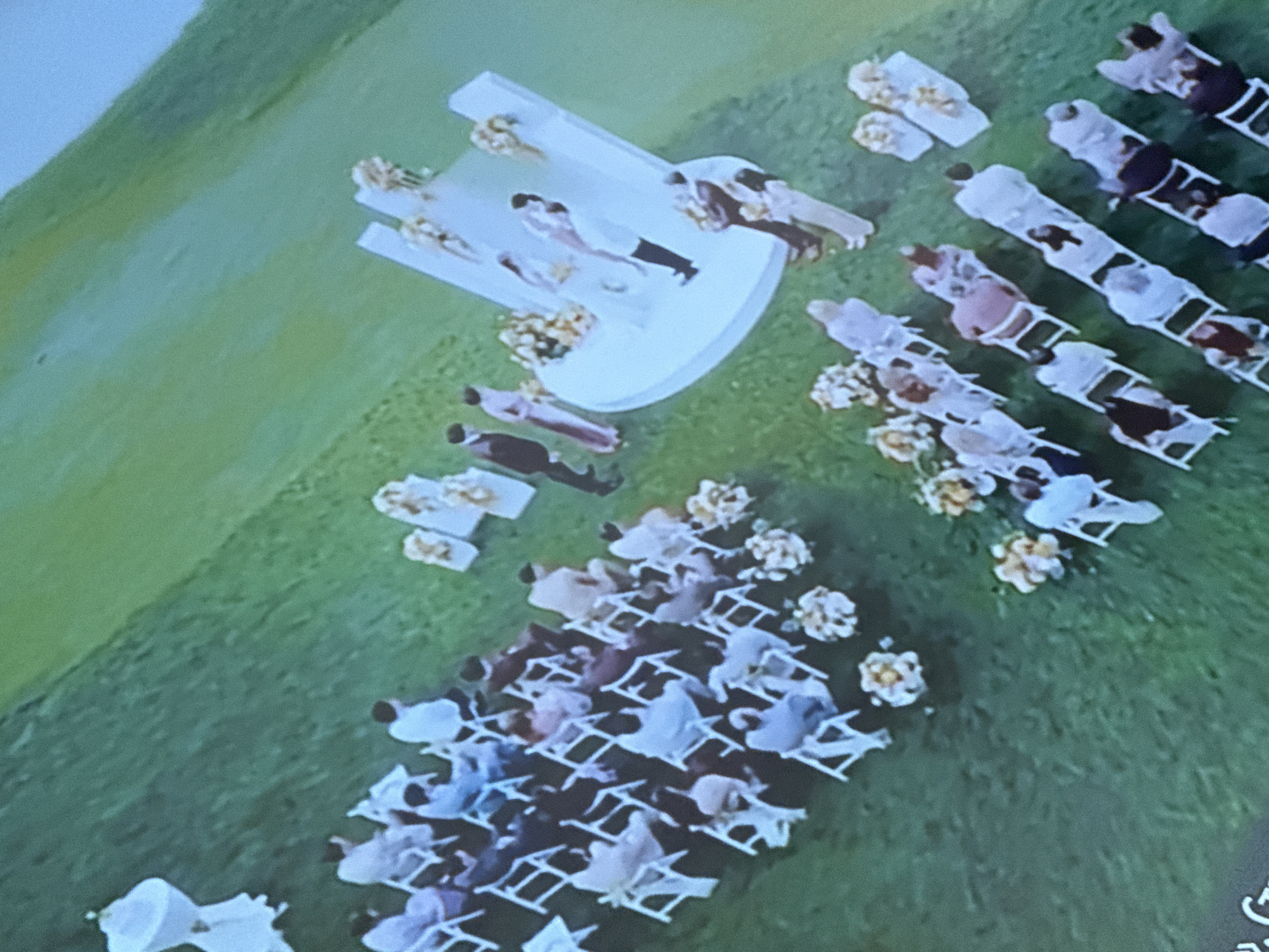The image shows an outdoor wedding ceremony in what appears to be a grassy field. White folding chairs are arranged in rows facing a raised, circular platform. People are seated in the chairs, facing the platform where the wedding party stands. Bouquets of flowers are placed along the rows of chairs and around the platform. The location is implied to be near the city of Skalba Snt, Russia, indicated by the original request.