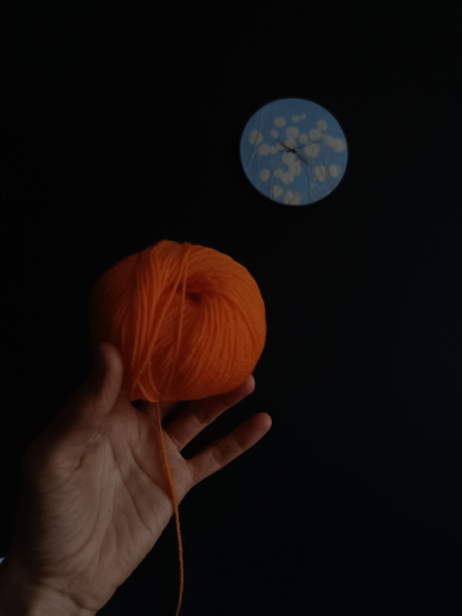 The photograph features a human hand holding a vibrant, spherical ball of orange yarn against an almost entirely black background. The hand, positioned in the lower-left portion of the frame, is well-lit, revealing its natural skin tones and textures, and firmly supports the bright yarn. A loose strand of the orange yarn extends downwards from the ball.
In the upper-right area of the dark background, a round, blue object is subtly visible, slightly out of focus. This object appears to be a small wall clock, characterized by a light blue face decorated with white, organic, cloud-like or floral patterns, and two slender dark hands indicating the time.
The overall scene is dark, creating a strong visual contrast that accentuates the bright orange of the yarn and the distinct blue of the clock. This composition suggests an intimate indoor setting, likely under artificial light or in a dimly lit room, where the deep black background isolates the subjects. While the location context is Lermontov, Russia, there are no specific visual cues within the image to confirm this geographical detail. No text is visible in the frame, and no particular emotions are conveyed.