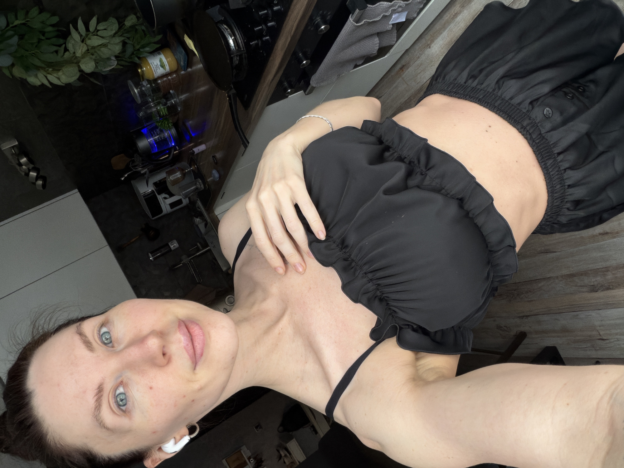 A selfie captures a woman with fair skin, blue eyes, and dark hair, likely in her 20s or 30s, looking directly at the camera with a neutral or slightly pleasant expression. She is wearing a black, ruffled top with thin straps and a black, elasticated skirt or shorts, exposing her midriff. A white earbud is visible in her left ear, and a silver braided bracelet is on her right wrist.
The background suggests an indoor setting, specifically a kitchen in Perm, Russia. On a dark wooden countertop, a black stovetop with a pan is visible, alongside a grey towel, and various items including a yellow bottle with a label showing "MACARONI PASTA" and green leaf graphics. Several clear glasses or bottles, one illuminated with a blue light, are also on the counter next to a kitchen appliance that resembles a coffee machine or blender. Green leafy plants are placed on a shelf above the counter. The lighting is diffused, and no external weather or strong natural light cues are present. The scene depicts a casual moment.