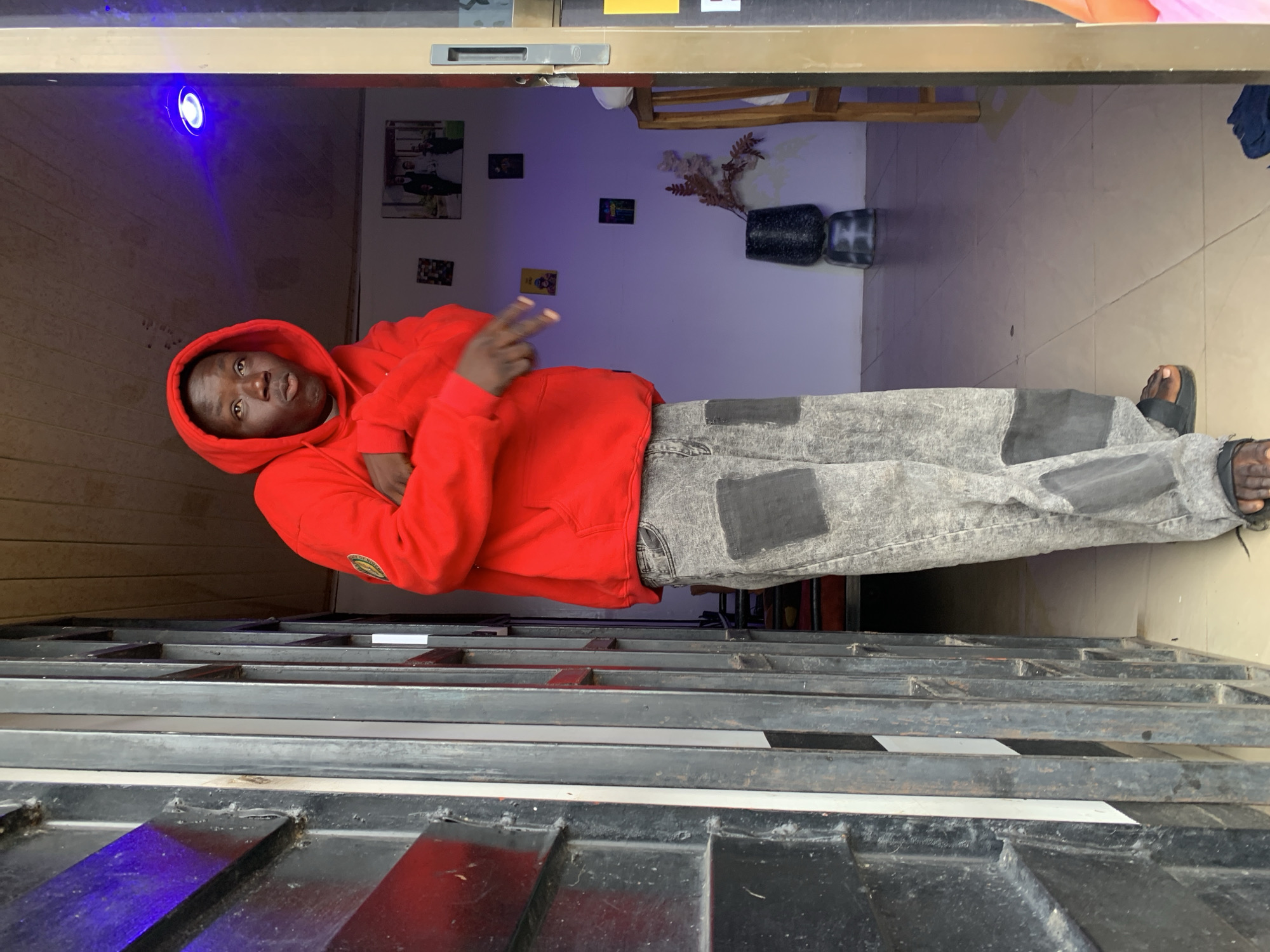 This image captures a male individual standing near an entrance, asserted to be in Jos, Nigeria.
The subject is a young male with a dark complexion, wearing a bright red hooded sweatshirt with the hood up. A circular yellow, green, and black patch is visible on his left sleeve. He is also wearing light grey, acid-washed or distressed wide-leg jeans with prominent dark grey rectangular patches on the thighs and knees, and black sandals on his feet. He is positioned slightly turned to his right, with his head looking directly towards the camera. His mouth is slightly open, and his right hand is raised with two fingers extended in a 'V' gesture. His left arm is bent across his chest.
The immediate foreground to the left is dominated by dark, multi-barred metal security gating or a door frame, partially obscuring the left side of the scene. A white vertical stripe is visible on one of the inner bars of this gate structure.
The background reveals an indoor setting with white walls and a light-colored tiled floor. A blue LED light fixture is mounted on the ceiling in the upper right, casting a distinct blue hue. Several framed pictures or artworks adorn the wall: a larger central image depicts a group including what appears to be a wedding couple, surrounded by smaller, colorful pictures. On the floor to the right, a dark, textured vase holding dried plant material rests on a smaller, similar base. A light-colored wooden chair or stool is partially visible to the right of the vase, and some dark fabric is on the floor in the bottom right corner. Part of a light-colored door frame with a dark handle is visible on the far right, suggesting an adjacent room or an exit. The ceiling appears light-colored, possibly painted wood.