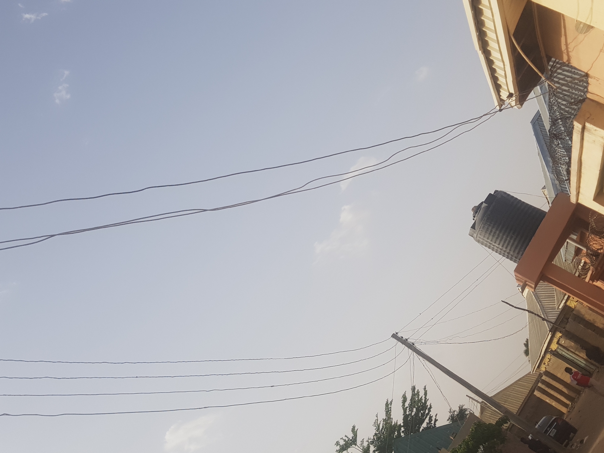 The image depicts a daytime scene in what appears to be a residential area of Dabawa, Nigeria. The sky is predominantly light blue with few clouds. Electrical wires crisscross the sky. Buildings of various styles and colors are visible, including structures with corrugated metal roofing. A large, black water tank stands elevated on a peach-colored platform attached to a building. A utility pole leans slightly, supporting more electrical wires. Green trees and vegetation are at the bottom of the frame. A black car is parked in front of one of the buildings, and a person wearing red clothing can be seen near the car.