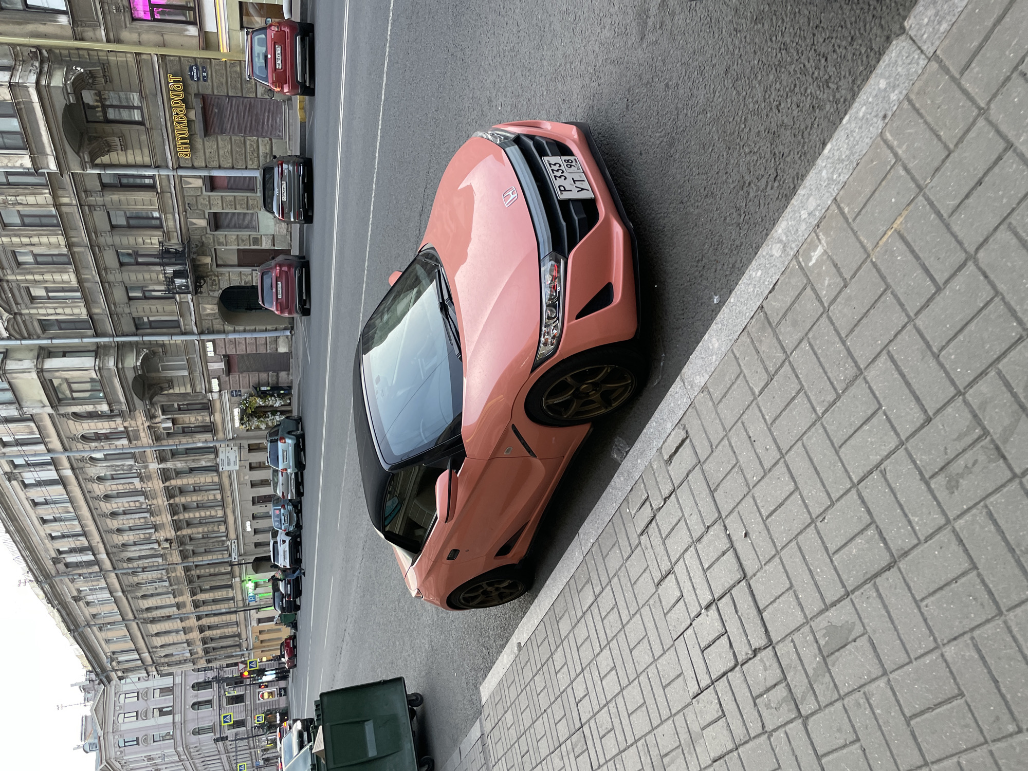 The image depicts a vibrant salmon pink Honda S660 convertible, featuring a black soft top and dark bronze multi-spoke alloy wheels, parked curb-side on a paved sidewalk. The car's front license plate reads "P 333 YT 98," indicating registration in Saint Petersburg, Russia. The sidewalk consists of grey rectangular concrete pavers laid in a herringbone pattern, with a granite curb separating it from the asphalt road. A green plastic wheeled trash bin is visible further down the sidewalk on the left.
The street is a multi-lane thoroughfare with double white lines marking the center. Several other vehicles are parked on the opposite side of the street, including a dark red SUV, a dark grey Audi A5 Sportback, and another red SUV. A person is visible walking on the far sidewalk near the parked cars.
The background features a continuous row of multi-story, historic buildings, characteristic of 19th and early 20th-century European architecture, with ornate facades, arched windows, and decorative balconies. Storefronts occupy the ground level of these buildings. A prominent yellow sign on one building reads "АНТИКВАРІАТ" (ANTIQUARIAT), indicating an antique shop, while another sign further down is partially visible as "АВТОЭКСПЕРТ" (AUTOEXPERT). Based on the architecture and these specific signs, the scene is located on **Liteyny Prospekt in Saint Petersburg, Russia**, specifically looking south-east from the sidewalk, with the "АНТИКВАРІАТ" shop located at Liteyny Prospekt 26. The sky appears overcast, suggesting diffused daylight.
