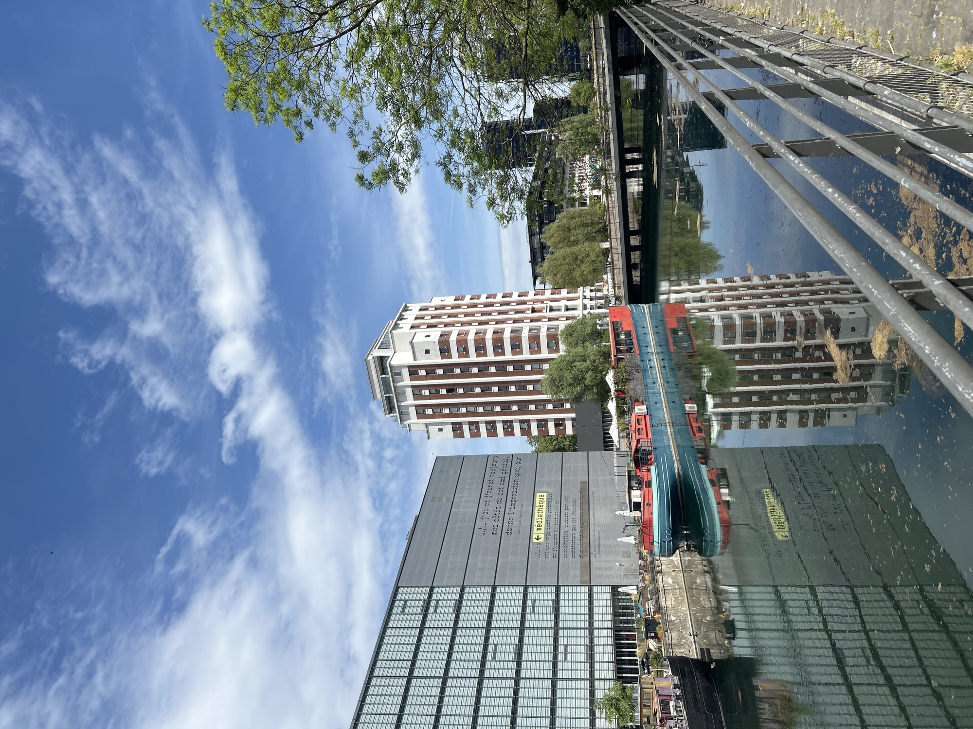 The image is a street-level view in Strasbourg, France, depicting architectural reflections in a canal or river. The sky is blue with scattered clouds, visible both in the sky and reflected in the water. Trees are present at the top of the frame, and the scene includes a railing along the canal. Several modern buildings are visible, including a high-rise with a red and white facade and another structure with reflective windows and an inscription in French. A barge is docked along the canal. The overall scene conveys a sense of urban life blended with natural elements in a European city.