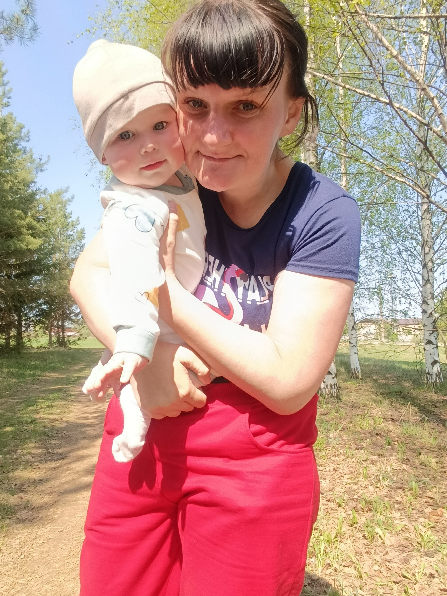 A woman in a navy blue t-shirt and red pants holds a baby wearing a beige hat and white onesie. They are outside, in an area with trees and greenery. A dirt path is visible on the left. The location is near Фоки, Russia.