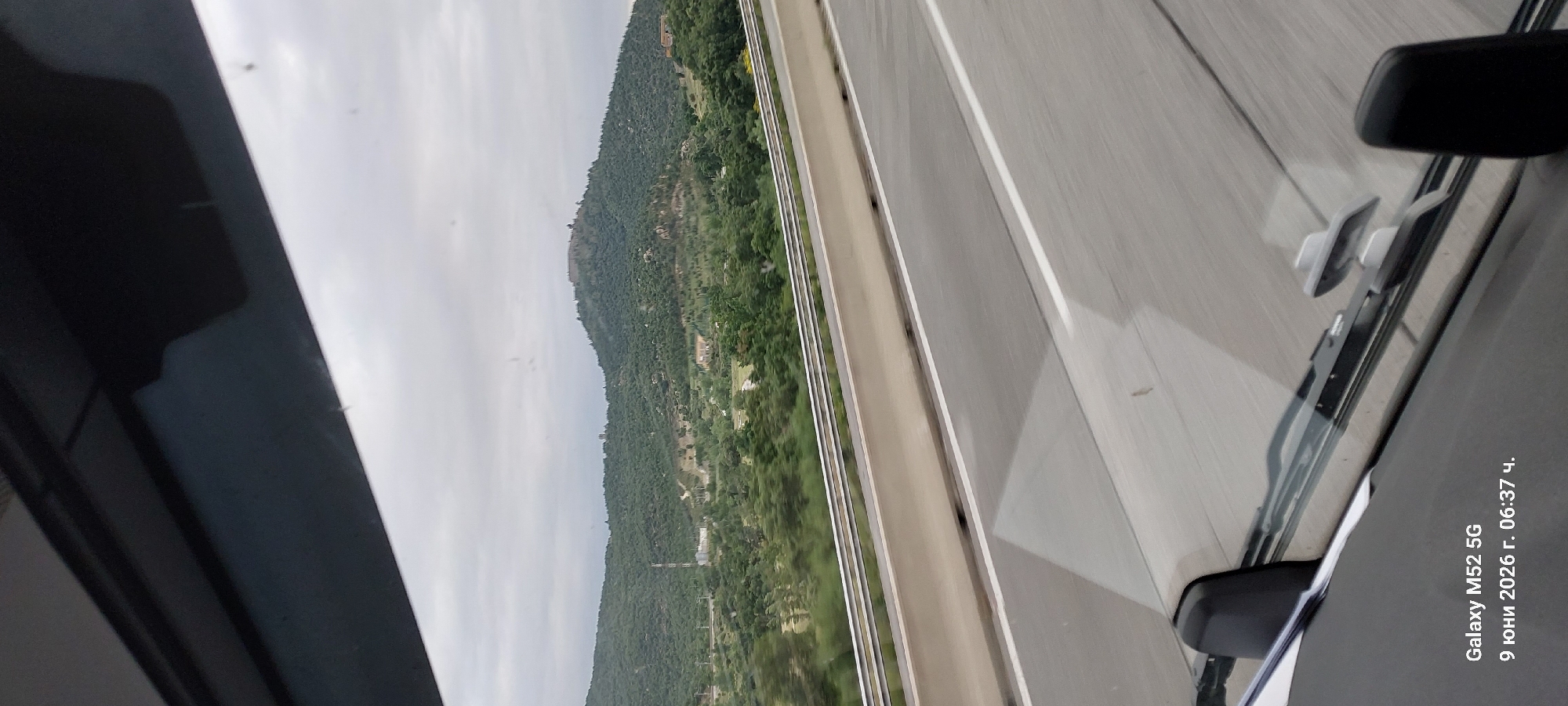 The image shows a view from inside a vehicle, looking out at a hilly landscape under an overcast sky. The foreground features the vehicle's dashboard, including a windshield wiper and part of the car's interior. The middle ground consists of a highway with concrete barriers and lanes. Beyond the highway, the landscape is covered with dense green vegetation, with scattered buildings and a communication tower visible on a distant hill. At the very top of the furthest hill, a structure resembling a fortification or ruins is discernible. The bottom left corner of the image displays text indicating "Galaxy M52 5G" and a date and time: "9 юни 2026 г. 06:37 ч." This date corresponds to June 9, 2026, at 6:37 AM. While the image itself does not explicitly show the city of La Jonquera, Spain, it depicts a roadside view consistent with the mountainous terrain surrounding such a location, likely captured during a journey.