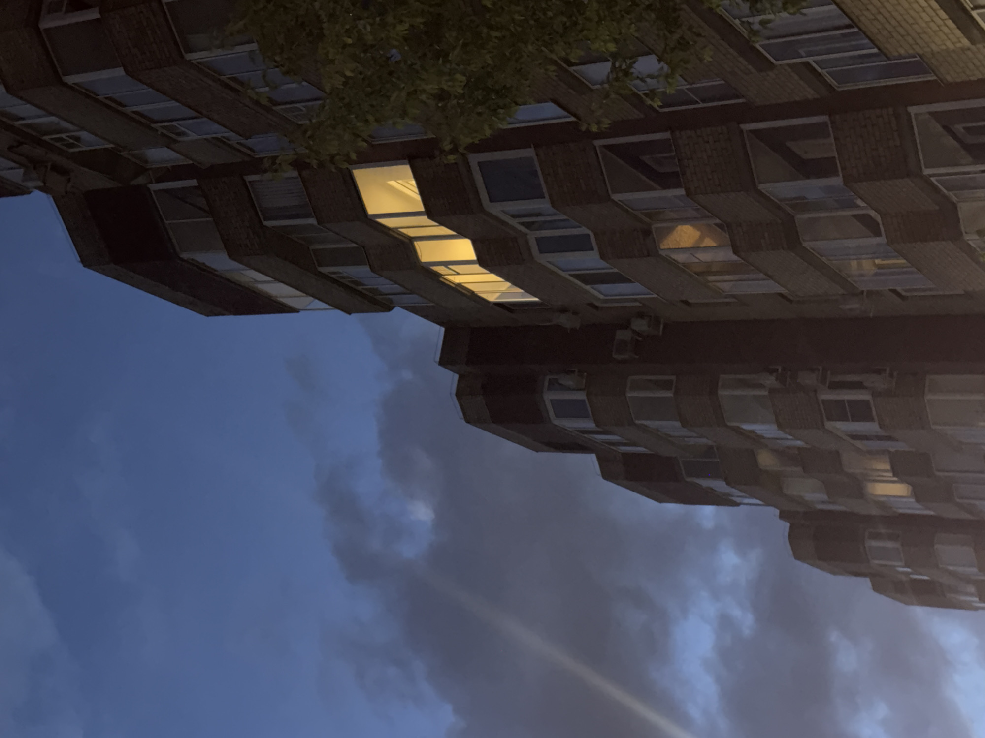 The image shows a multi-story brick building, likely an apartment complex, at dusk or dawn. The building is characterized by its staggered balcony or window arrangement. A few windows are lit, emanating a warm yellow light. The sky is a mix of dark blue and cloudy gray. The building's location is not discernible as Perm, Russia, from this image alone. There are no visible people, actions, or interactions.