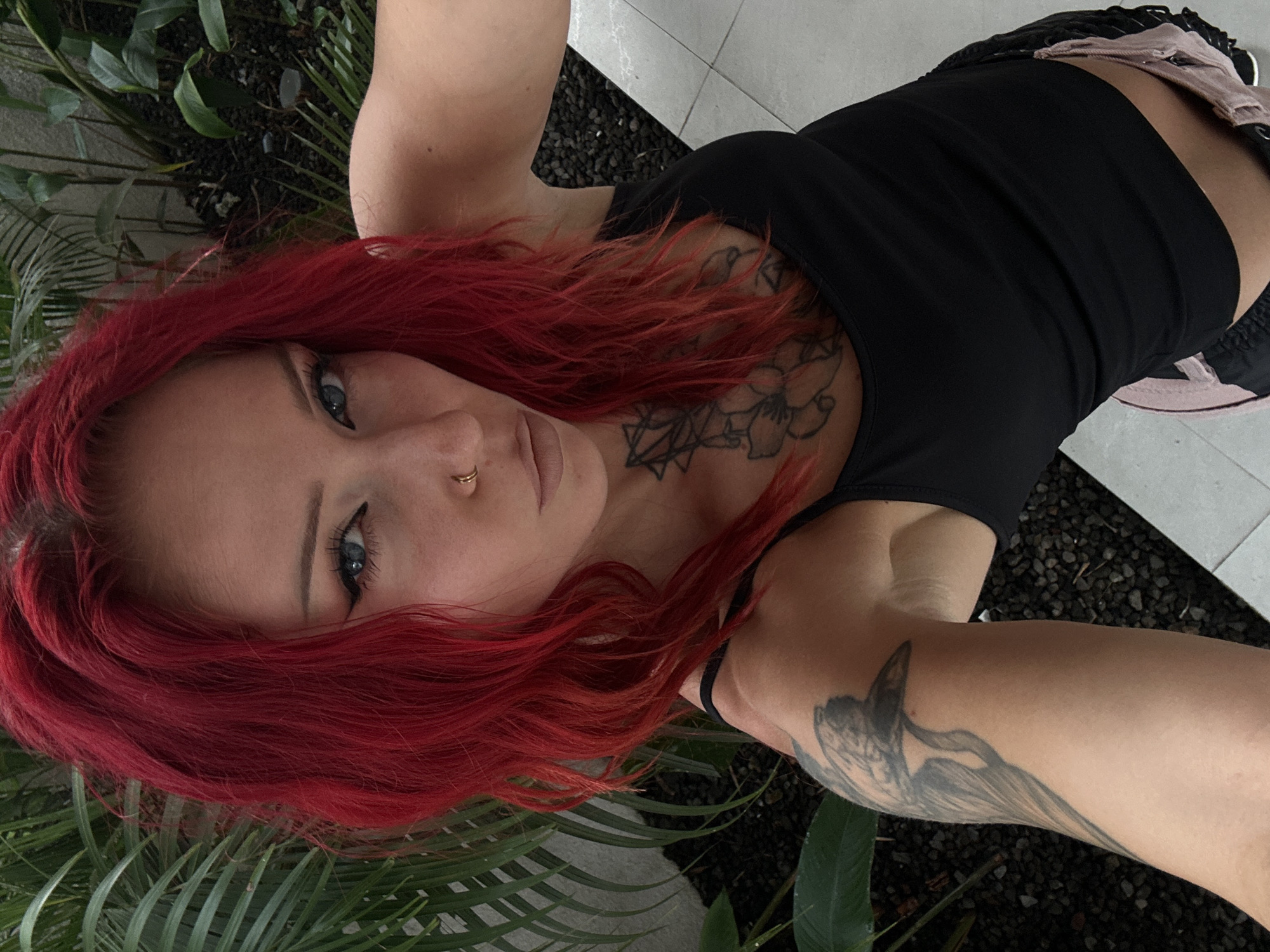 A woman with vibrant red hair, blue eyes, and a nose ring is taking a selfie outdoors in North Kuta, Indonesia. She is wearing a black tank top, and has tattoos on her chest and arm. She is standing in front of lush green foliage and dark gravel. The ground is tiled.