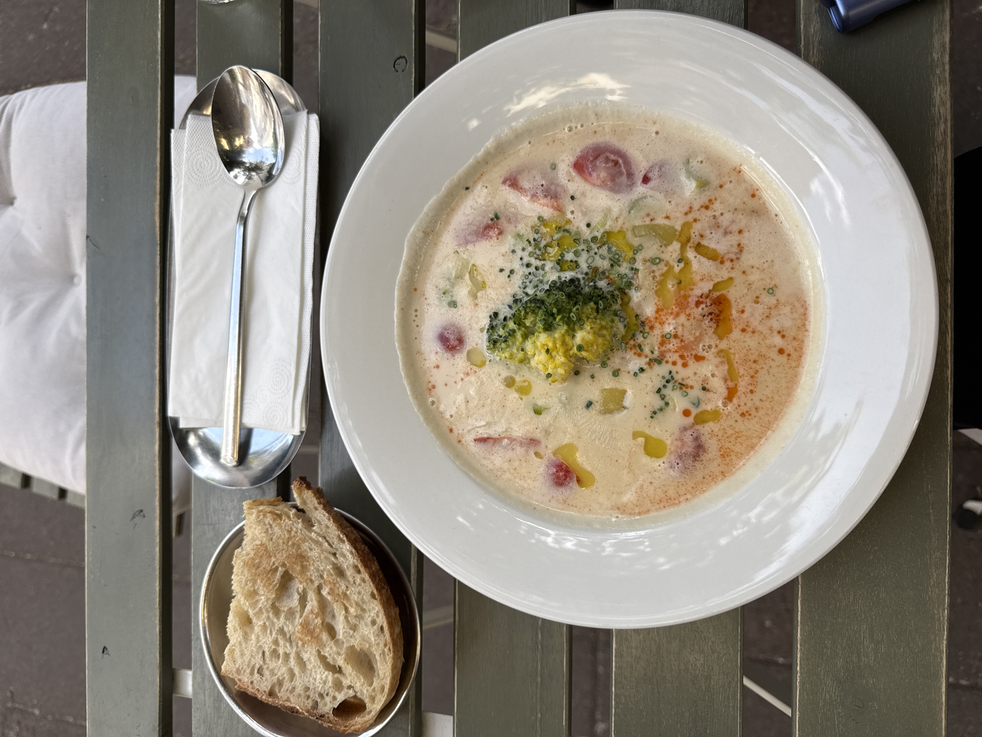 A top-down shot on a weathered green table shows a white bowl of creamy soup, garnished with a broccoli floret, diced vegetables, and oil droplets, alongside a metal container with a slice of bread, and another metal container holding a spoon and a napkin. The scene likely takes place at an outdoor dining area in Belgrade, Serbia.