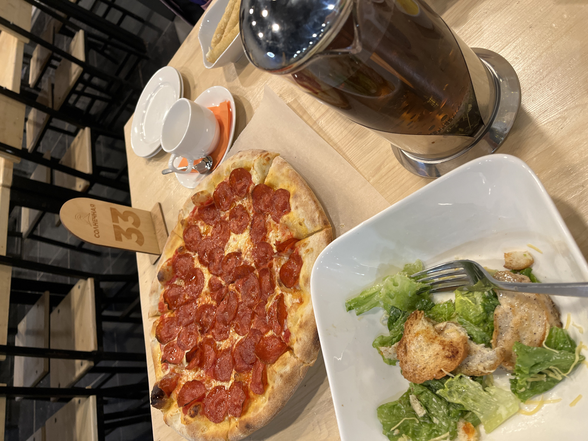 The image captures a dining scene, likely indoors, featuring a wooden table upon which various food items are arranged. Dominating the composition is a pepperoni pizza, cut into slices, placed on a sheet of parchment paper. Nearby is a white bowl containing a Caesar salad, with a fork resting on top. A glass teapot filled with tea sits adjacent to the salad. A small white cup and saucer, along with two white plates, are also visible on the table. Additionally, a small dish containing some sort of fried dough food is located behind the cup. A table number sign reads "33". From the prompt it is implied that the photo was taken in Paratunka, Russia. The presence of the pizza, salad, and tea suggests a casual meal setting. The chairs in the background suggest a restaurant or cafe environment.