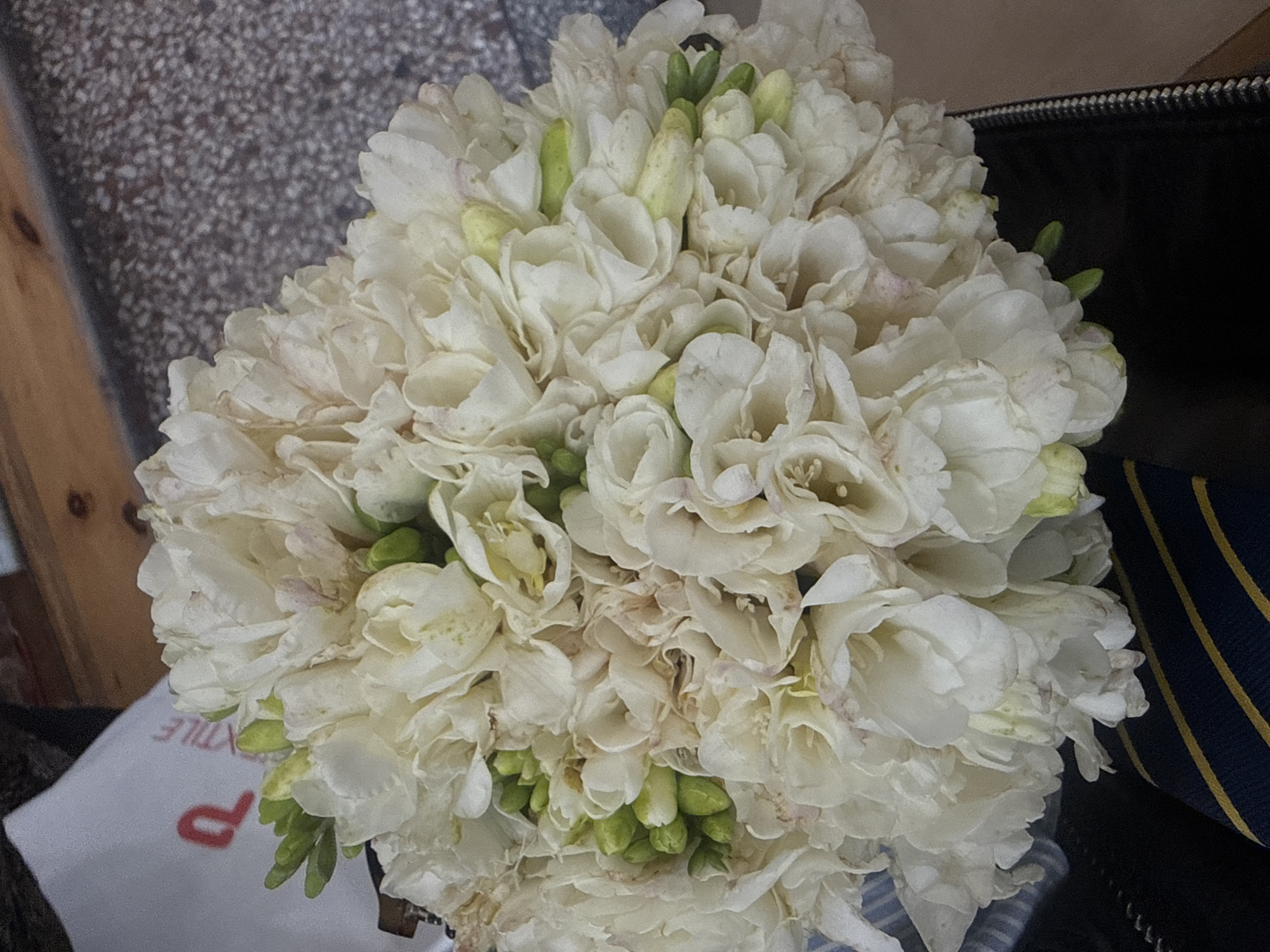 The image shows a close-up of a dense bouquet of white flowers, potentially freesias, with some green buds interspersed throughout. The bouquet appears to be resting on a surface alongside several items. To the left, there's a surface with a texture resembling speckled gravel or patterned fabric. Below the flowers, a partially visible white object with a red letter "P" and the word "TEXTILE" can be seen. To the right of the bouquet, there is a dark, shiny object that could be a bag or leather item, with a striped dark blue and gold object adjacent to it. Without additional context, the location in Yerevan, Armenia cannot be confirmed, and no people, actions, or interactions are observed.