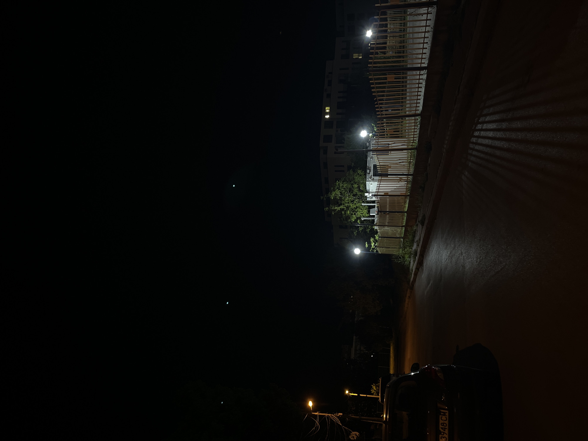 The image depicts a quiet nighttime street scene in Sofia, Bulgaria. The setting features a paved road or path running along the right side, bordered by a fence and buildings.
Multiple streetlights illuminate parts of the scene. On the right, two bright white streetlights cast light on a slatted fence or barrier, creating prominent, elongated parallel shadows that stretch across the dark ground. Further down and to the left, another streetlight emits a warmer, yellow glow. In the upper left, a modern building with illuminated windows is partially visible against the dark sky. A dark-colored car is parked on the side of the road in the bottom right, with its rear visible. The car's license plate clearly displays "3948 CM," which is a Bulgarian plate code for Sofia, confirming the location.
The ground appears dark and potentially reflective, possibly from dampness. The overall atmosphere is still and devoid of human activity. The time of day is clearly night, with the sole sources of illumination being artificial lights.