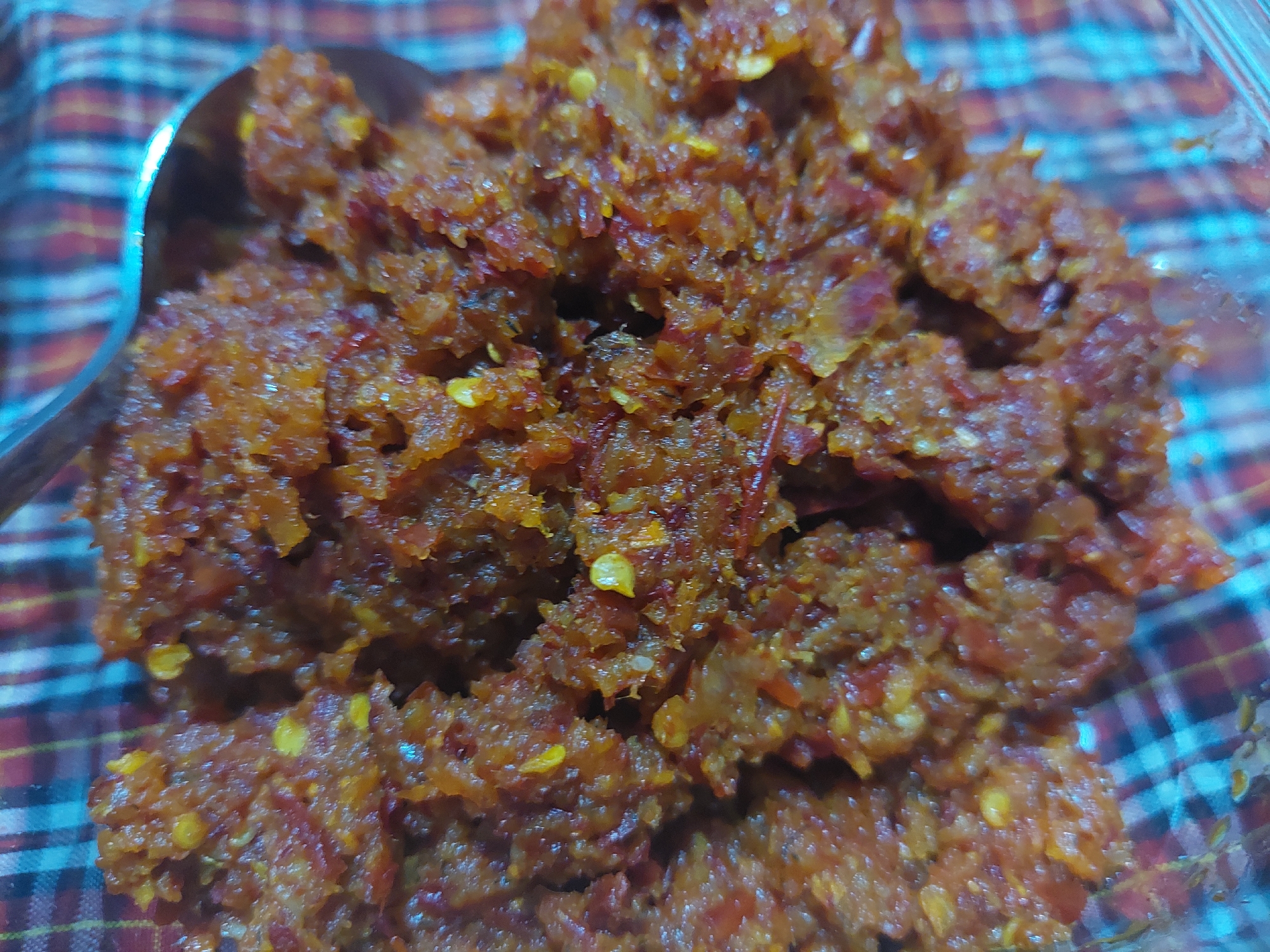 A close-up, high-angle shot shows a serving of sambal, a spicy chili paste, in a clear container. The sambal is a coarse, reddish-brown mixture with visible chili seeds and flakes, suggesting it is freshly made or of a chunky texture. A portion of a silver spoon is visible on the left side of the frame, partially submerged in the sambal, indicating it is either being served or has just been used. The background is a blurred checkered fabric with stripes of red, white, and blue, providing a vibrant contrast to the food. The image is well-lit, with the light source creating subtle highlights on the sambal and the spoon. The scene is static, with no discernible action or people present. The overall impression is of a food item prepared and ready for consumption, likely in a domestic or casual setting within Jelutung, Indonesia, given the context provided.