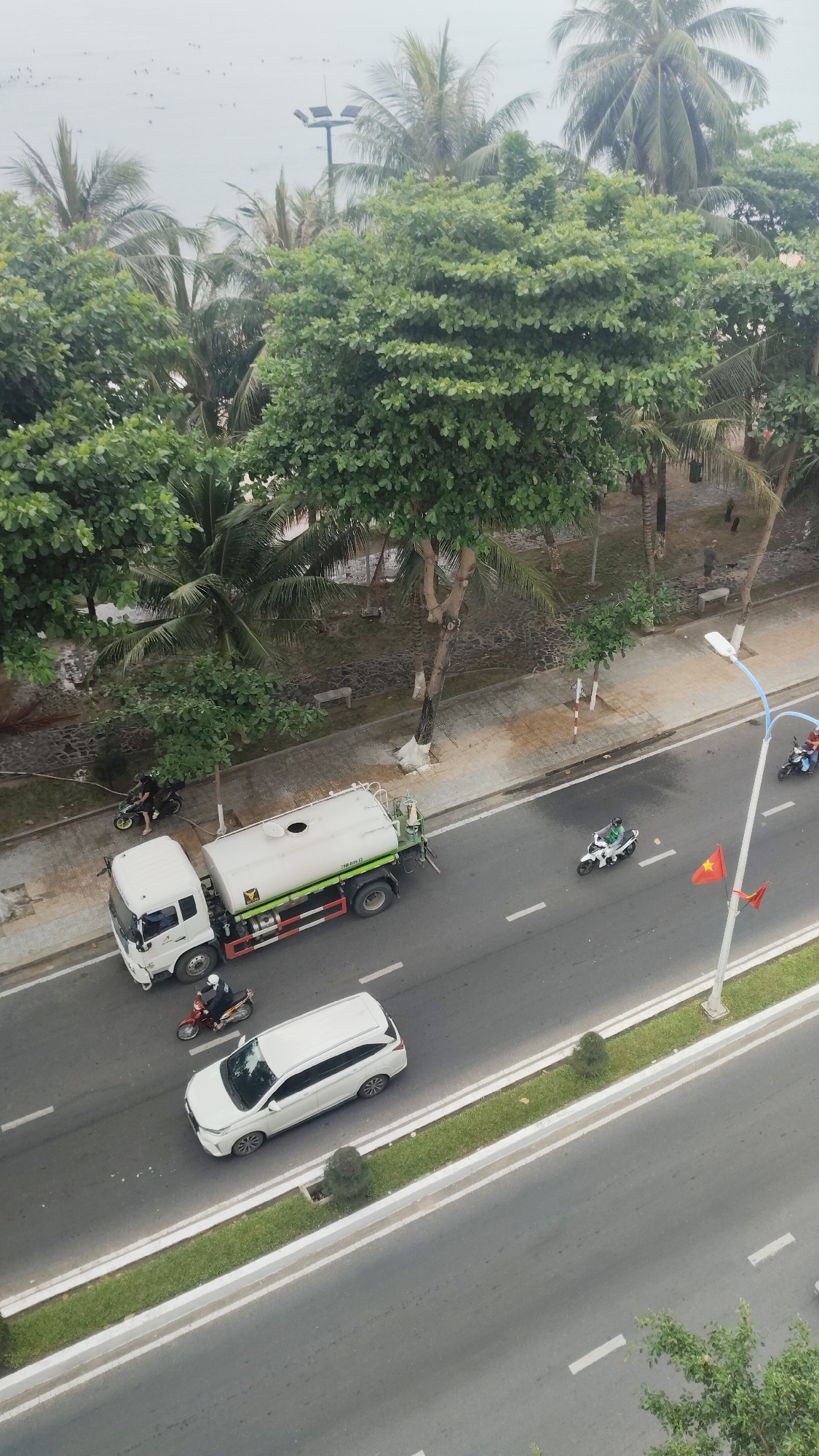 The image shows a street scene in Bac Nha Trang Ward, Vietnam, taken from an elevated perspective. A white water truck is parked on the side of the road near the sidewalk. A white car is driving alongside it on the road. Two motorcycles are riding nearby the truck and the car. There are several people on motorcycles and on foot. A row of trees lines the street. There are palm trees and a beach in the background. A Vietnamese flag is mounted on a pole.