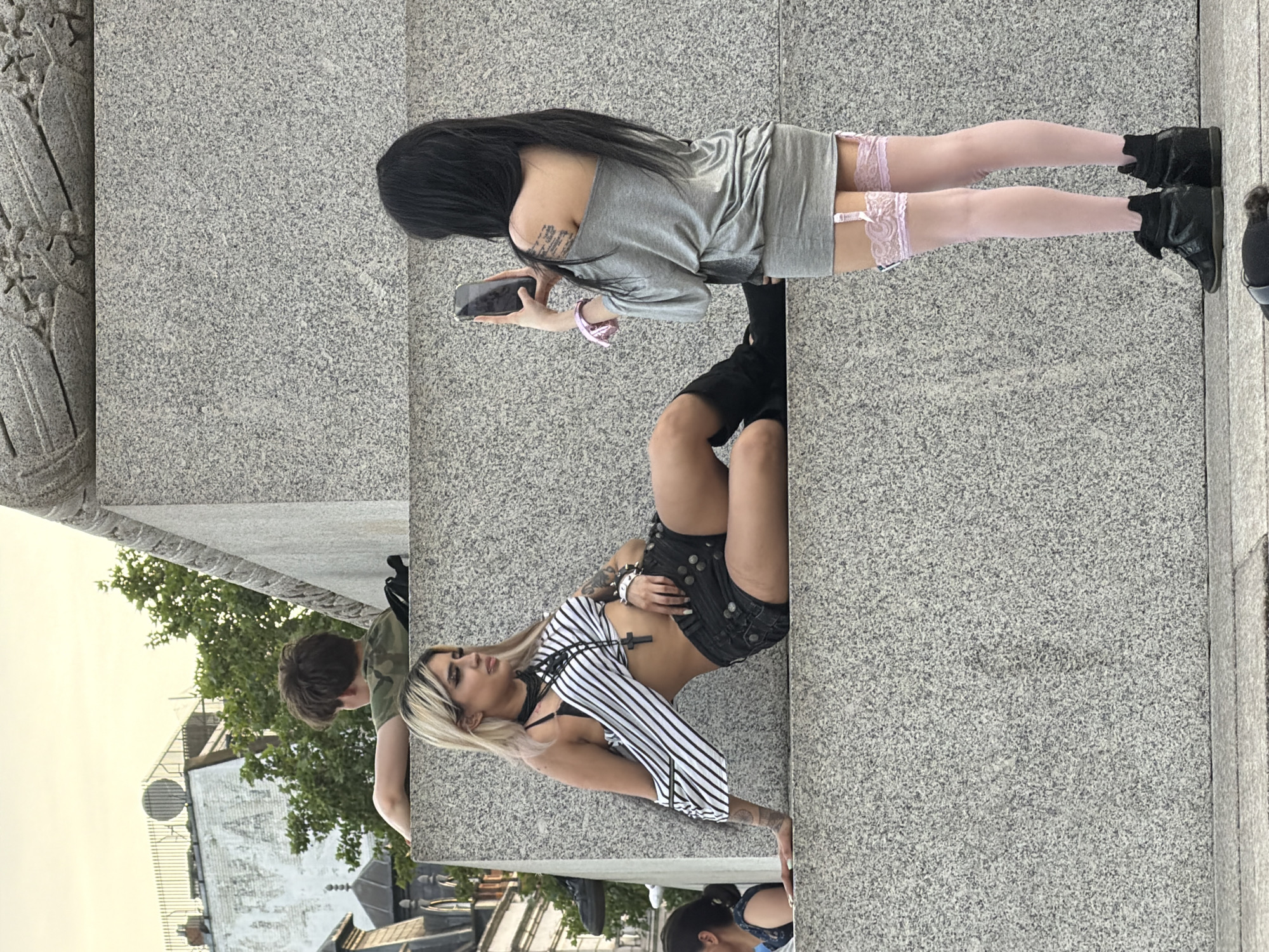 The photograph depicts an outdoor scene in London, United Kingdom. Two women and a man are positioned on a raised, textured grey surface which forms part of an architectural structure. One woman, with long black hair, is facing away from the camera and looking at a mobile phone. She is wearing a short grey dress, light pink thigh-high stockings, and black boots. The second woman is wearing a striped top, black shorts, and has blonde hair. The man is wearing a green camouflage shirt. They appear to be posing for a photo. The background includes buildings and greenery, suggesting an urban location. The lighting suggests daylight hours.