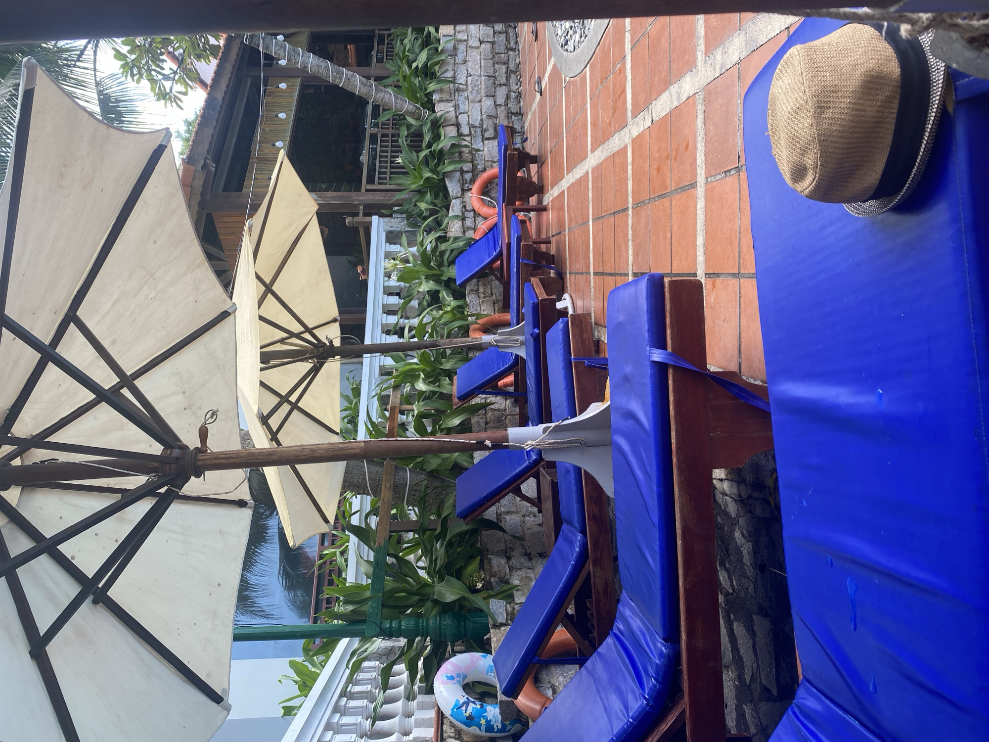 This image captures a tranquil outdoor leisure area, specifically located in Phu Thuy Ward, Vietnam. The scene features multiple wooden lounge chairs equipped with vibrant blue cushions, arranged on a reddish-brown tiled patio. In the immediate foreground, a light brown straw fedora with a dark band rests upon the cushion of the closest lounge chair, which, along with others, displays scattered water droplets. Overhead, several large, open, cream-colored square umbrellas, supported by wooden frames, provide shade. To the left, a stone wall supports lush green foliage and a white balustrade, with a blue and white inflatable ring resting on the wall. Further back, two standard orange ring buoys are visible near other lounge chairs. The mid-ground includes additional dense green plants along a textured stone retaining wall. In the background, wooden structures with reddish tiled roofs, consistent with resort architecture, are partially visible, connected by a string of bare bulb lights. A palm tree trunk, wrapped in white material, stands adjacent to these structures. On the right, black flip-flops are placed on the tiled ground near a curved, stone-edged section, possibly indicating the perimeter of a pool or water feature. The area is devoid of people, presenting an unoccupied and calm atmosphere under bright daylight.
