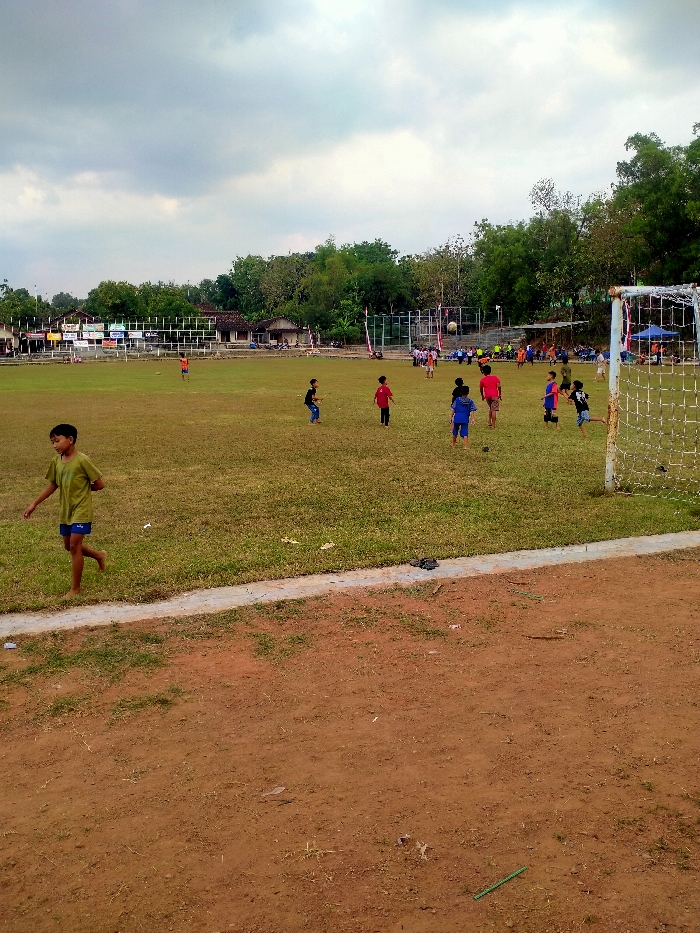 An outdoor scene captures a football field in Dawung, Indonesia, under an overcast sky. The foreground features a reddish-brown dirt area with sparse vegetation, bordered by a narrow concrete strip, beyond which lies the main playing field of dry, green grass with visible brown patches. Approximately 15 to 20 individuals, predominantly children, are dispersed across the field. One child, wearing a light green shirt and blue shorts, walks barefoot in the left foreground, moving right. Several groups of children are actively engaged in playing football in the mid-ground, with a white football visibly airborne near a cluster of players. A white metal football goal post with a net is partially visible on the far right. The background consists of a low, horizontal barrier, beyond which are single-story structures with various roofs and signs, followed by a dense line of trees with green foliage. Additional individuals are present in the background, some seated under a blue canopy structure.