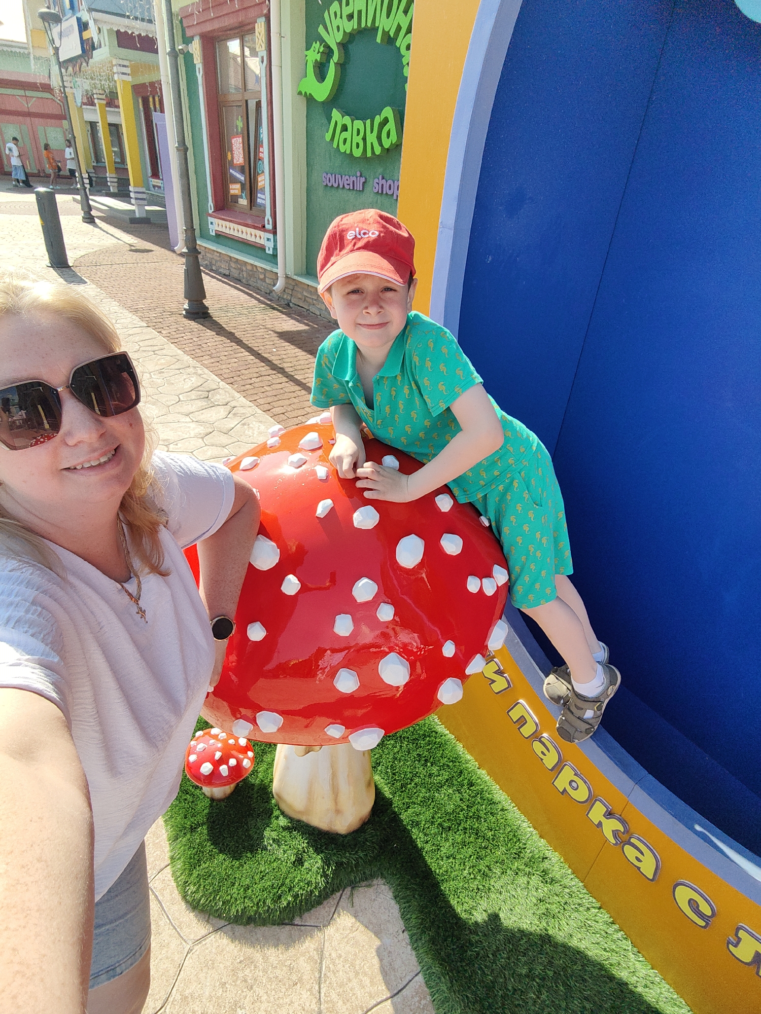 In a sun-drenched outdoor setting, a woman with blonde hair and sunglasses is taking a selfie. She's wearing a light pink t-shirt and grey shorts. Next to her, a young boy in a green shortsleeve shirt with a repeating seahorse pattern and a red cap with "elco" written on it is playfully perched on a large, red mushroom sculpture with white spots. The mushroom is on a patch of artificial green grass. In the background, a brightly colored row of buildings with intricate details lines a paved walkway. One building has green and yellow accents and text in Russian. The scene appears to be a lively amusement park or tourist area, likely in Sochi, Russia, given the context. The bright sunlight suggests it's a warm, clear day. The overall mood is cheerful and relaxed, with the boy's posture conveying a sense of fun and adventure. Visible text includes "лавка" and "souvenir shop" on one of the buildings, and partial Russian text in yellow lettering on a blue and yellow curved structure behind the mushroom.