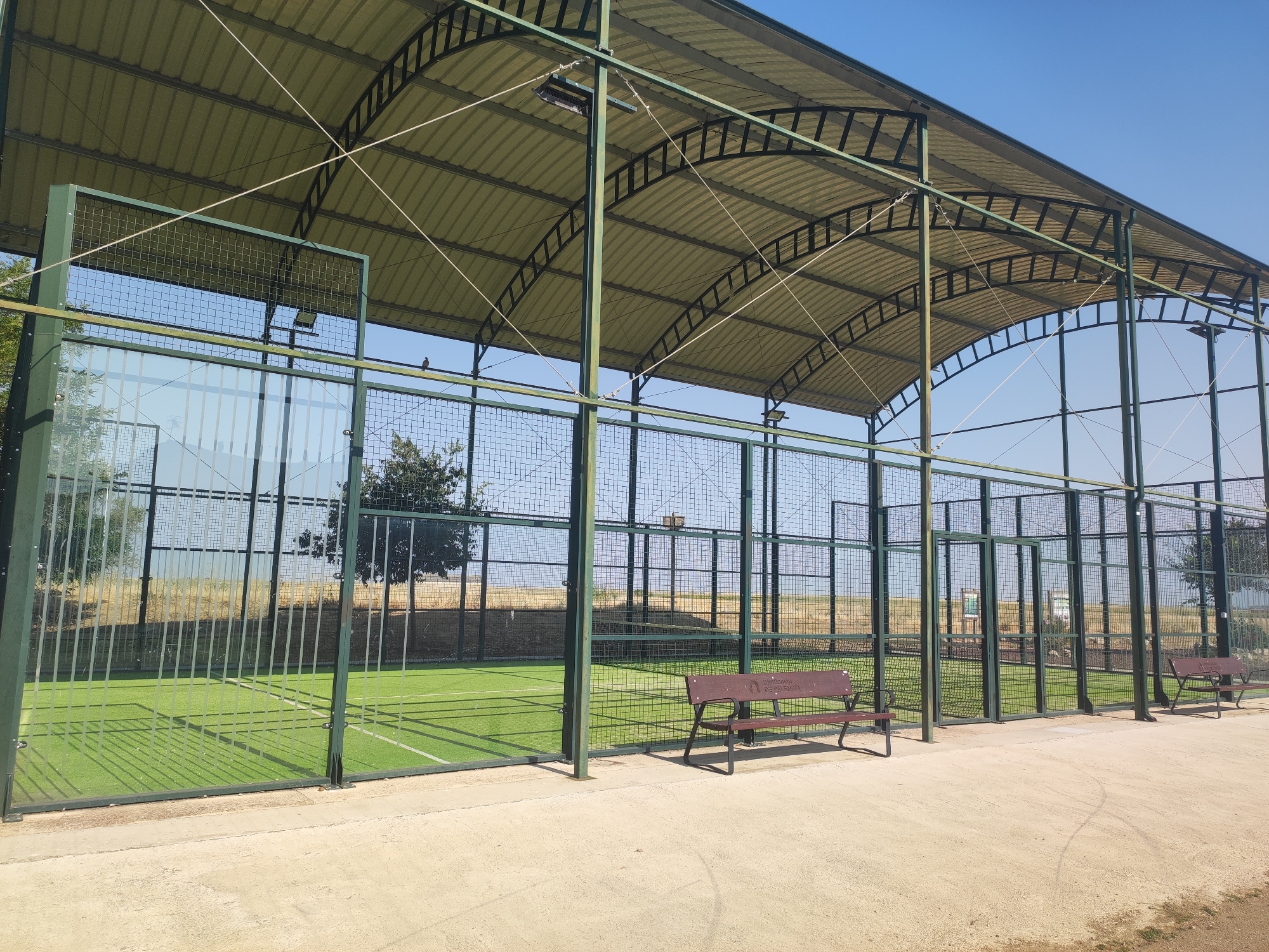 The image depicts an outdoor padel tennis court complex in Autillo de Campos, Spain. The courts are covered by a large, translucent, arched roof supported by a green metal framework. The courts themselves have a bright green artificial turf surface with white lines marking the boundaries. The perimeter of the courts is enclosed by a combination of green metal posts and wire mesh fencing, with sections of clear glass panels at the lower half of the back walls.
In the foreground, a concrete pathway runs alongside the courts. Two dark red metal benches are positioned on this pathway, facing the courts. One bench is closer to the camera and appears to have some text or a logo on its backrest. The environment surrounding the courts is dry and grassy, with some trees visible in the distance. The sky is clear and blue. The scene is static, with no people present. The lighting suggests it is daytime, with the sun casting shadows.
