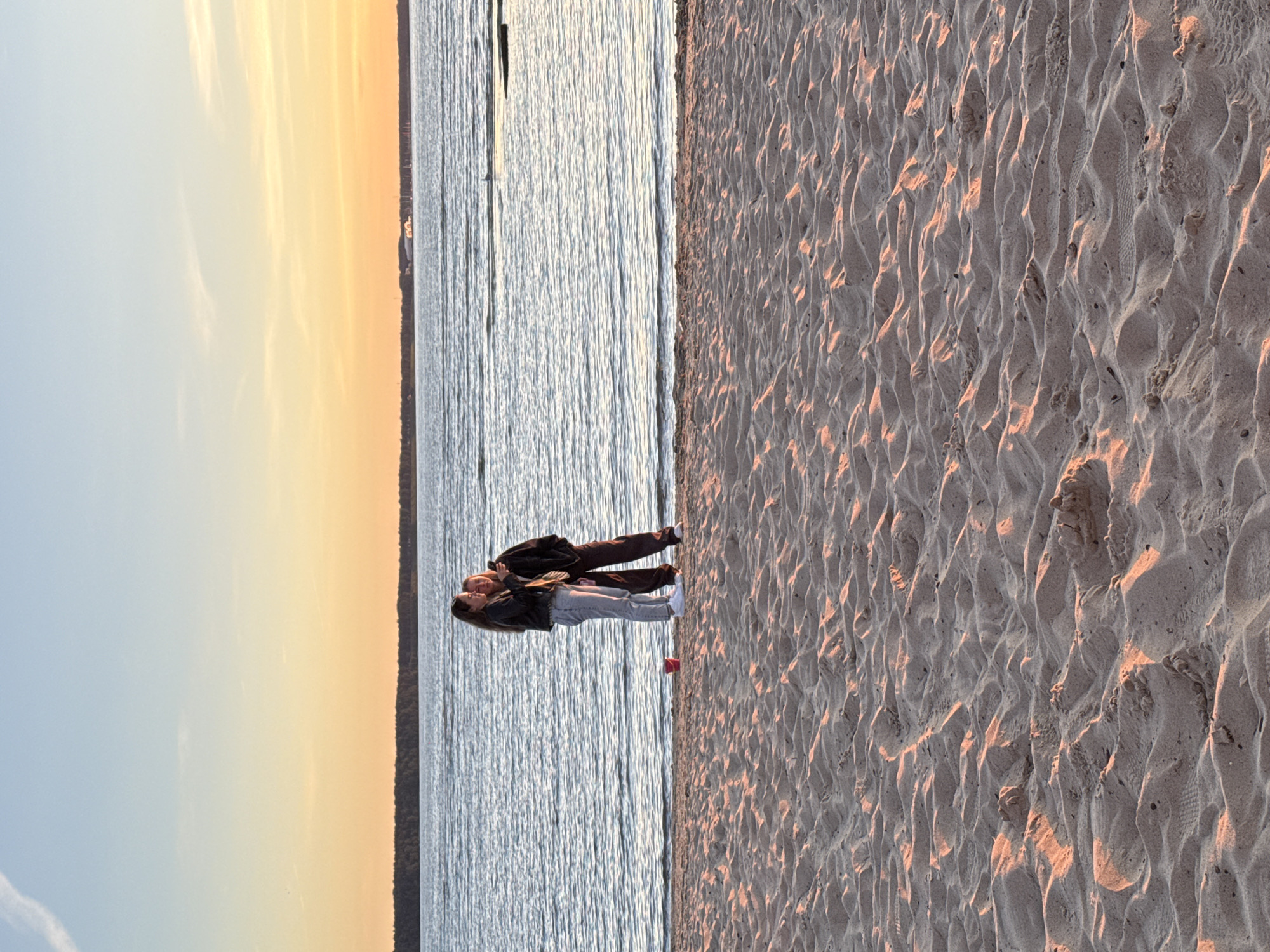 Two young females stand on a sandy beach by the water. The female on the left is wearing light gray jeans and a dark jacket. The female on the right is wearing dark pants and a dark jacket. Both are looking at a phone held by the female on the right. The water is calm with gentle ripples. In the background, a tree-lined horizon is visible with a few distant buildings and boats. The sky is a gradient of pale blue at the top, transitioning to a warm orange and yellow near the horizon, indicating either sunrise or sunset. The sand in the foreground is textured with ripples and small mounds, lit by warm, golden light. The scene appears to be set in Tallinn, Estonia, along the Baltic Sea coast.