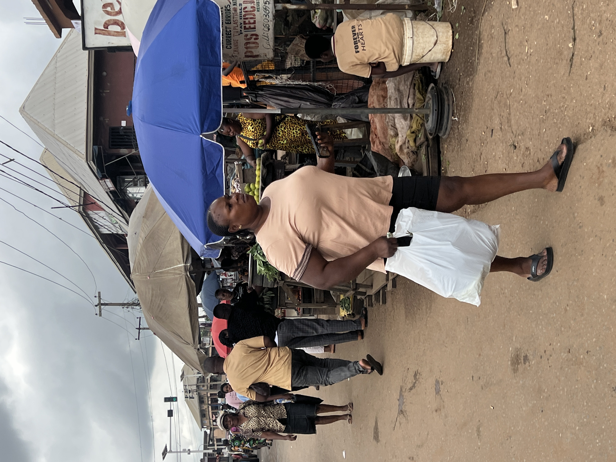 The image captures a street scene in Nkpor, Nigeria, with a focus on the vibrant market atmosphere. A woman in a light peach t-shirt and black shorts walks towards the viewer, carrying a large white plastic bag. Her expression is neutral as she navigates the busy street. Behind her, other individuals are visible, including a woman in a leopard-print dress and another person in a tan t-shirt with "FOREVER HEARTS" printed on it. Stalls and umbrellas are set up, indicative of a market setting. A blue umbrella is prominently featured, shielding some of the market activities. The ground appears to be a dirt or asphalt road, with scattered debris. Overhead, electrical wires crisscross the sky, suggesting an urban or semi-urban environment. The sky is overcast, hinting at a cloudy or possibly rainy day. The overall impression is of a typical day in a bustling Nigerian market, with people engaged in commerce and daily life. Visible text includes market signage with phrases like "CORRECT ERRORS," "POSEEDC," and a phone number. The scene conveys a sense of everyday activity and community engagement within the market.