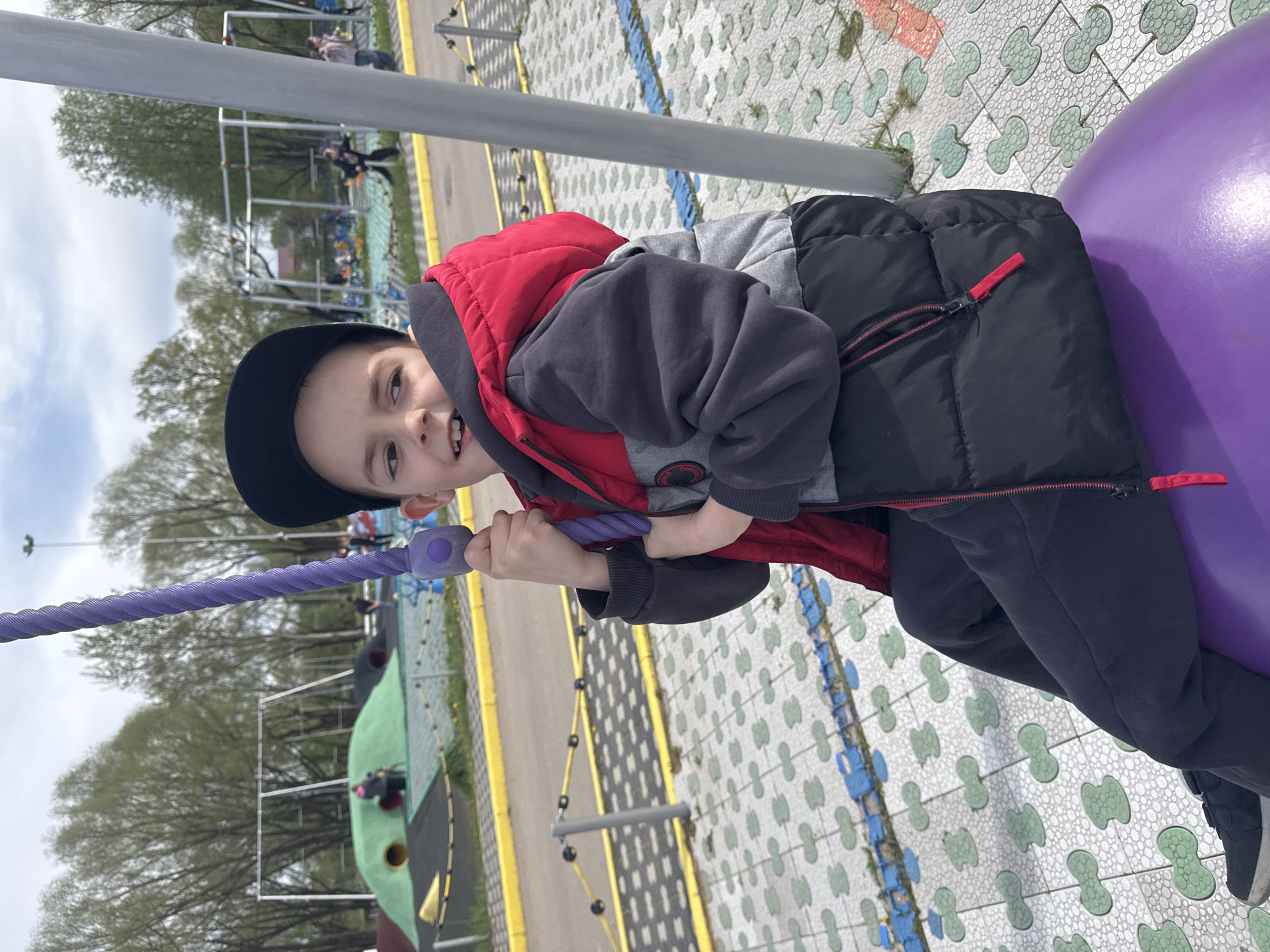 A young boy wearing a black cap, a red and gray hoodie, and a black jacket is playing on playground equipment in Синьково, Russia. He is holding onto a purple rope attached to a large purple ball. The playground has a patterned surface and other playground structures can be seen in the background. Trees are visible beyond the playground.