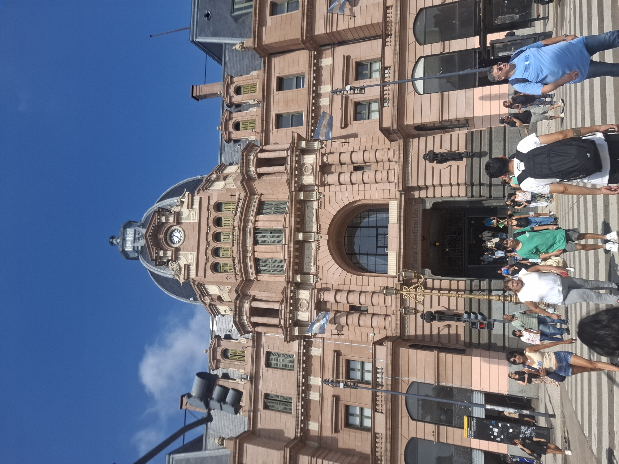 The image shows a street scene in Buenos Aires, Argentina, featuring the Plaza Constitución train station, specifically its iconic pink façade and clock tower. Several people are seen walking on the pedestrian crossing in front of the building, some wearing casual summer clothing. Street lights and traffic signals are visible in the foreground, while the clear blue sky forms the backdrop for the station's architecture. The building's entrance is visible, with people entering and exiting.