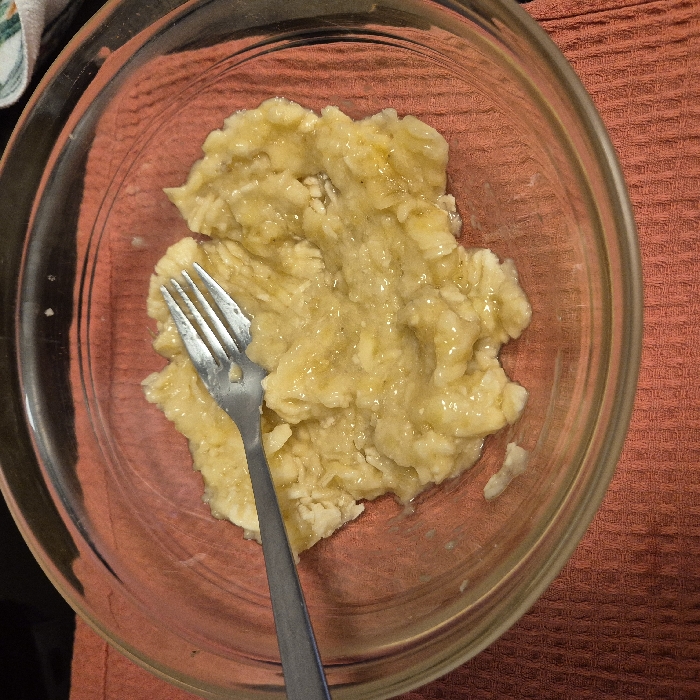 A clear glass bowl contains a quantity of mashed ripe bananas, exhibiting a lumpy, yellowish-brown consistency. A silver-toned metal eating fork is partially submerged, resting with its tines in the mashed fruit and its handle extending towards the upper left. The bowl sits on a reddish-orange, textured fabric surface, likely a kitchen towel or mat, which features a prominent waffle weave pattern. The background beyond the immediate setup is dark and indistinct. The specific location within the city of Strasbourg, France, cannot be determined from the visual information provided.