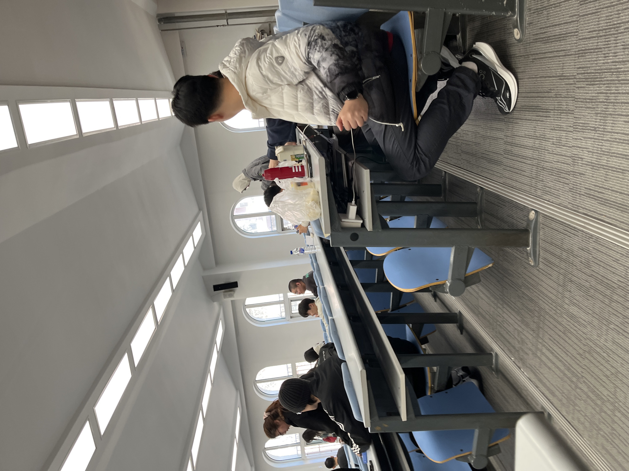 Location: Likely a lecture hall in Changchun City, China.
Scene: A row of students sits at desks in a lecture hall. The desks are attached, with blue seats. Students are facing forward. Some appear to be using electronic devices. A student in the foreground wears a white jacket with gray accents, black pants, and black and white sneakers. The room is well-lit with a row of rectangular windows. The floor has a gray carpet.