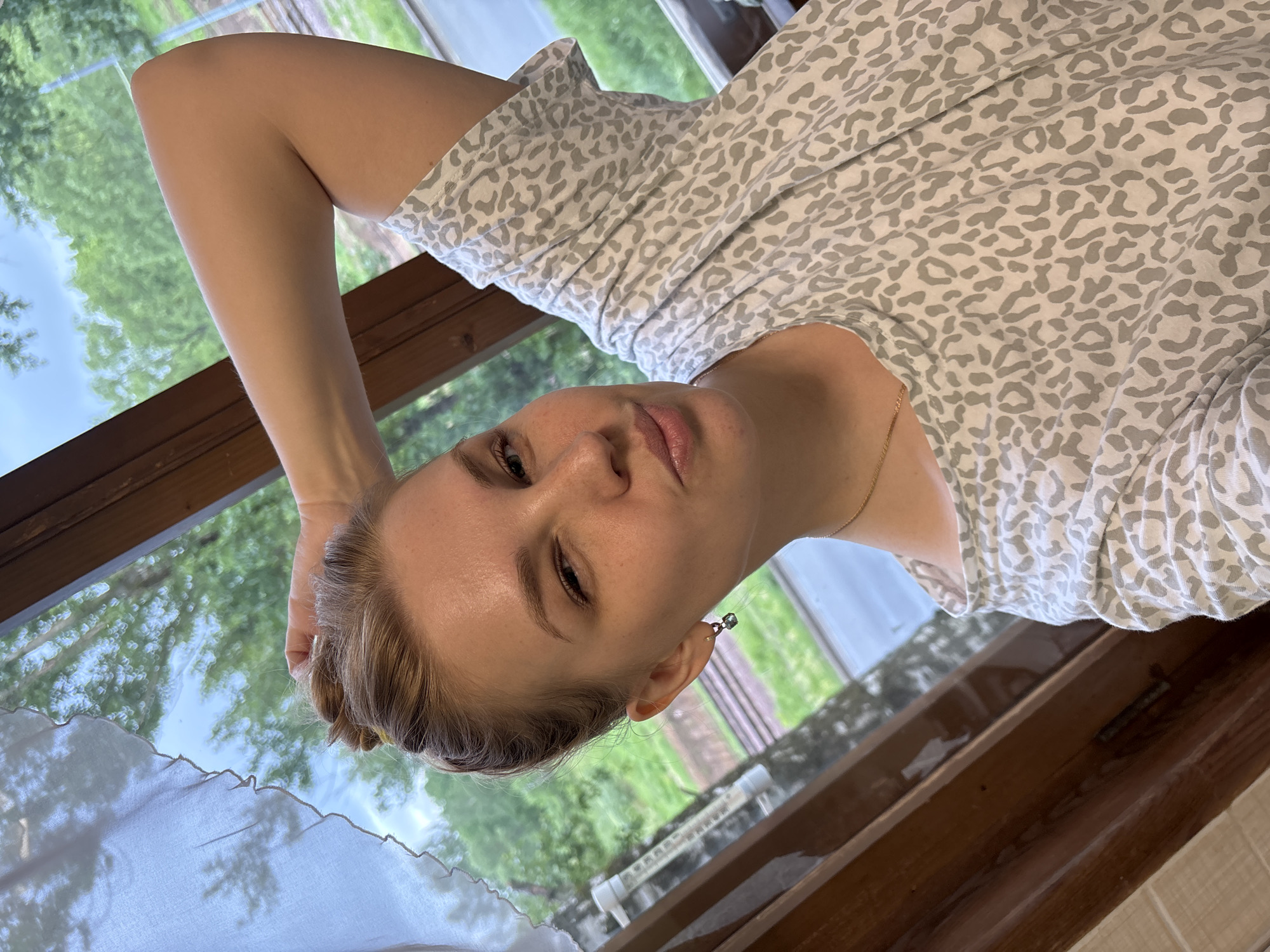 A fair-skinned woman is indoors, likely in a house, with a view of trees and sky visible through a window behind her. The woman is wearing a light-colored shirt with a grey animal print pattern. Her hair is blonde and appears to be tied back. She has an earring in her left ear. Her left arm is raised and her hand is positioned behind her head. There is a white curtain visible in the foreground near the bottom left of the frame. The window frame is made of dark wood. The location is uncertain, but I have no information that this is in the city of Rusanovo, Russia.