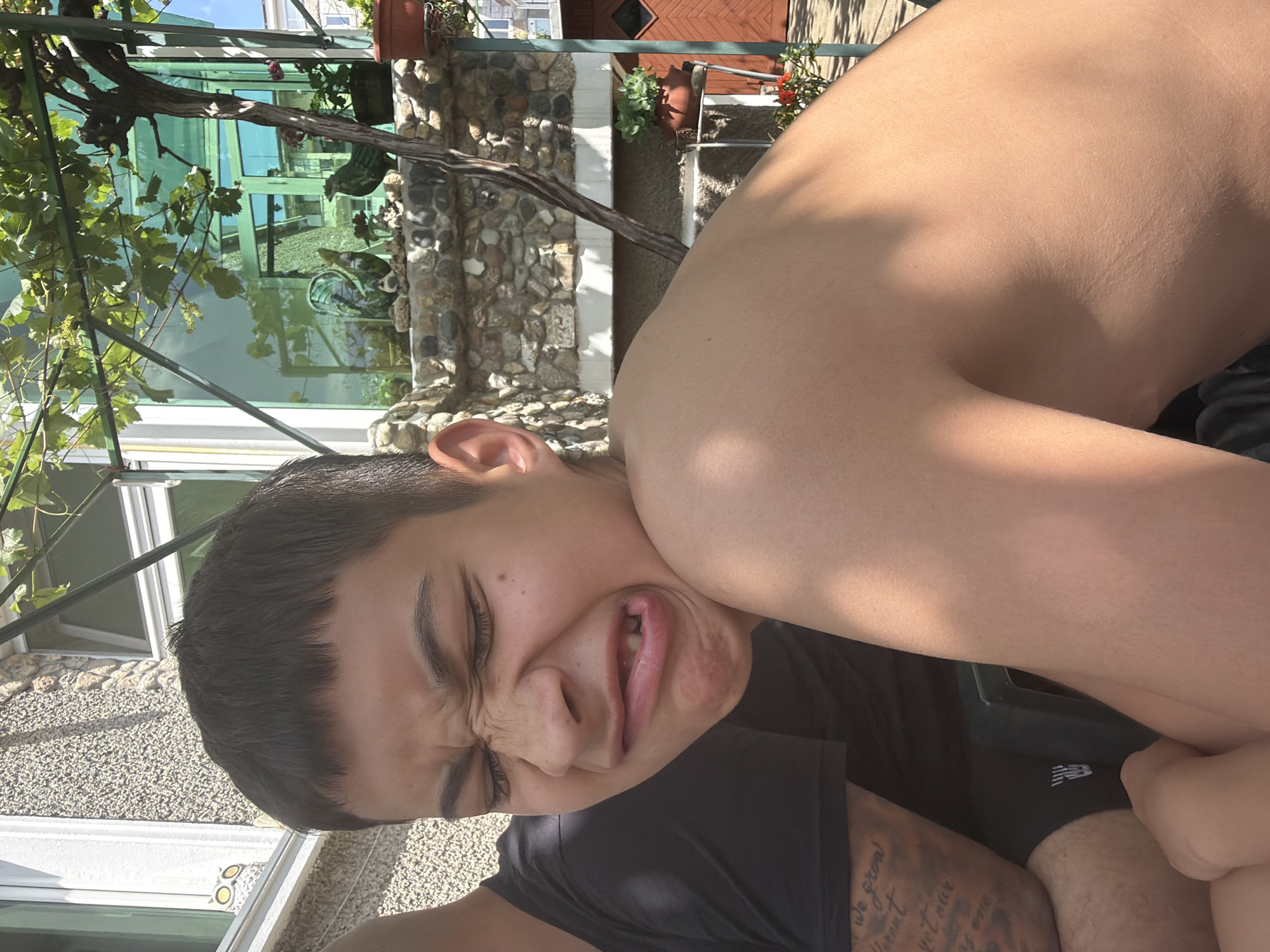 The image is a brightly lit, close-up, and rotated portrait of a young individual, likely male, captured in an outdoor setting. The subject, positioned diagonally across the frame, is making a humorous and exaggerated facial expression, with eyes squinted tightly, a crinkled nose, and an open mouth revealing teeth and tongue, conveying a playful mood. They have short, dark hair.
The person is wearing a black t-shirt. Visible on their arm or leg is a tattoo with fragmented text, including phrases like "We grow," "different," "yet our," and "as one." Another dark garment, possibly shorts or swimwear, with a white New Balance logo, is visible in the lower right portion of the image.
The background suggests an outdoor space on a sunny day, with strong shadows visible on the person's bare shoulder and the surroundings. Elements of the setting include a green, vine-covered structure, possibly a pergola or trellis, with lush green leaves and a gnarled vine. Beyond this, there are stone walls or decorative fencing. Architectural details include large glass windows or panels reflecting the environment, and a wooden structure with horizontal slats in the upper background. Potted plants, one in a terracotta pot and another with red blossoms, are also visible, contributing to a vibrant outdoor ambiance. The time of day appears to be mid-day or early afternoon due to the intense sunlight.