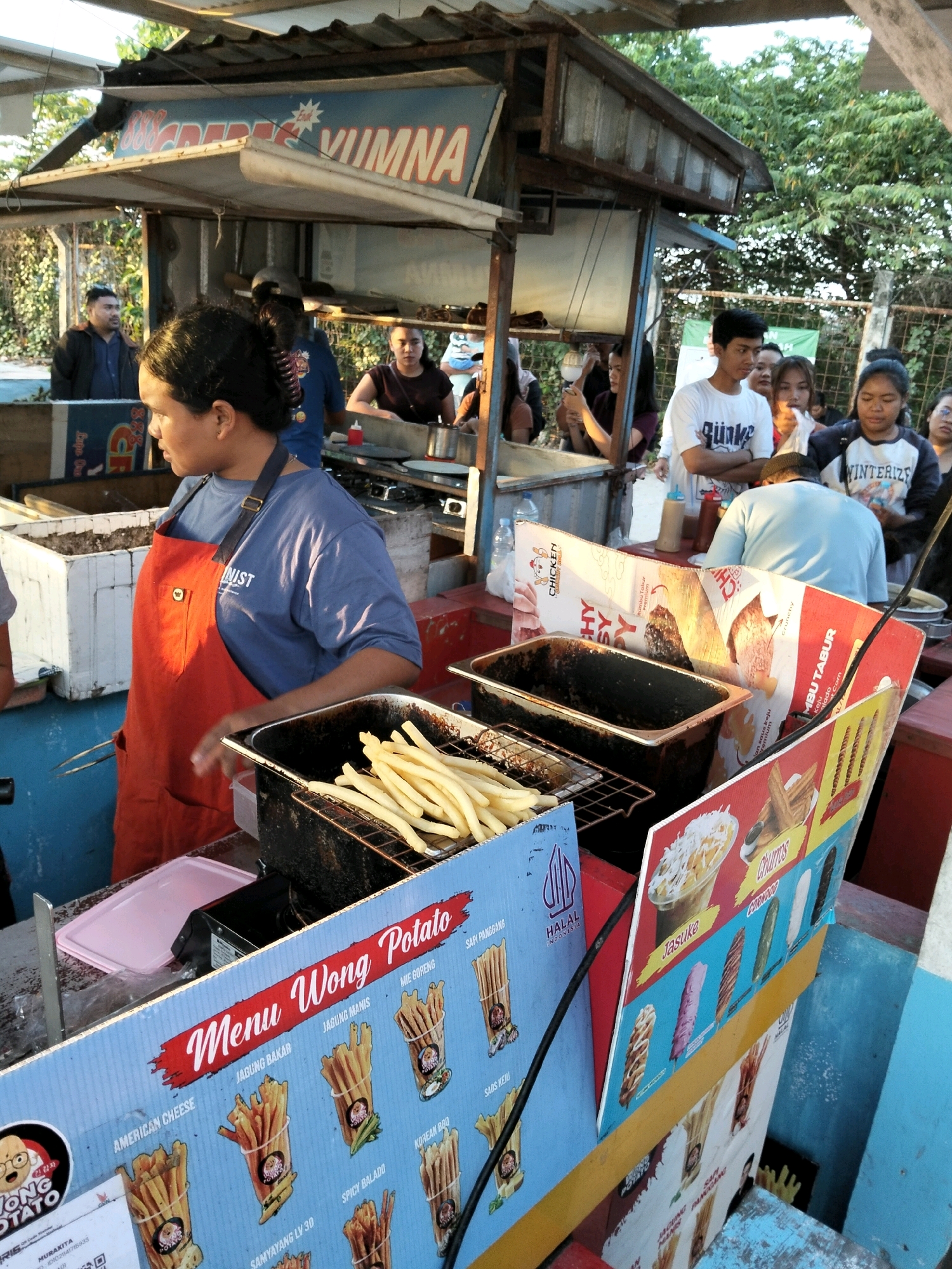 The image shows an outdoor food stall in Kuta, Indonesia. A woman wearing an orange apron and a blue t-shirt is working behind the counter, preparing food. She appears to be serving french fries, which are visible in a metal basket. Several other people are present, some are customers and others appear to be staff. The stall has signs advertising various food items, including "Wong Potato" and "Churros". The background shows trees and foliage, suggesting an outdoor setting. The overall impression is a busy street food vendor operation.