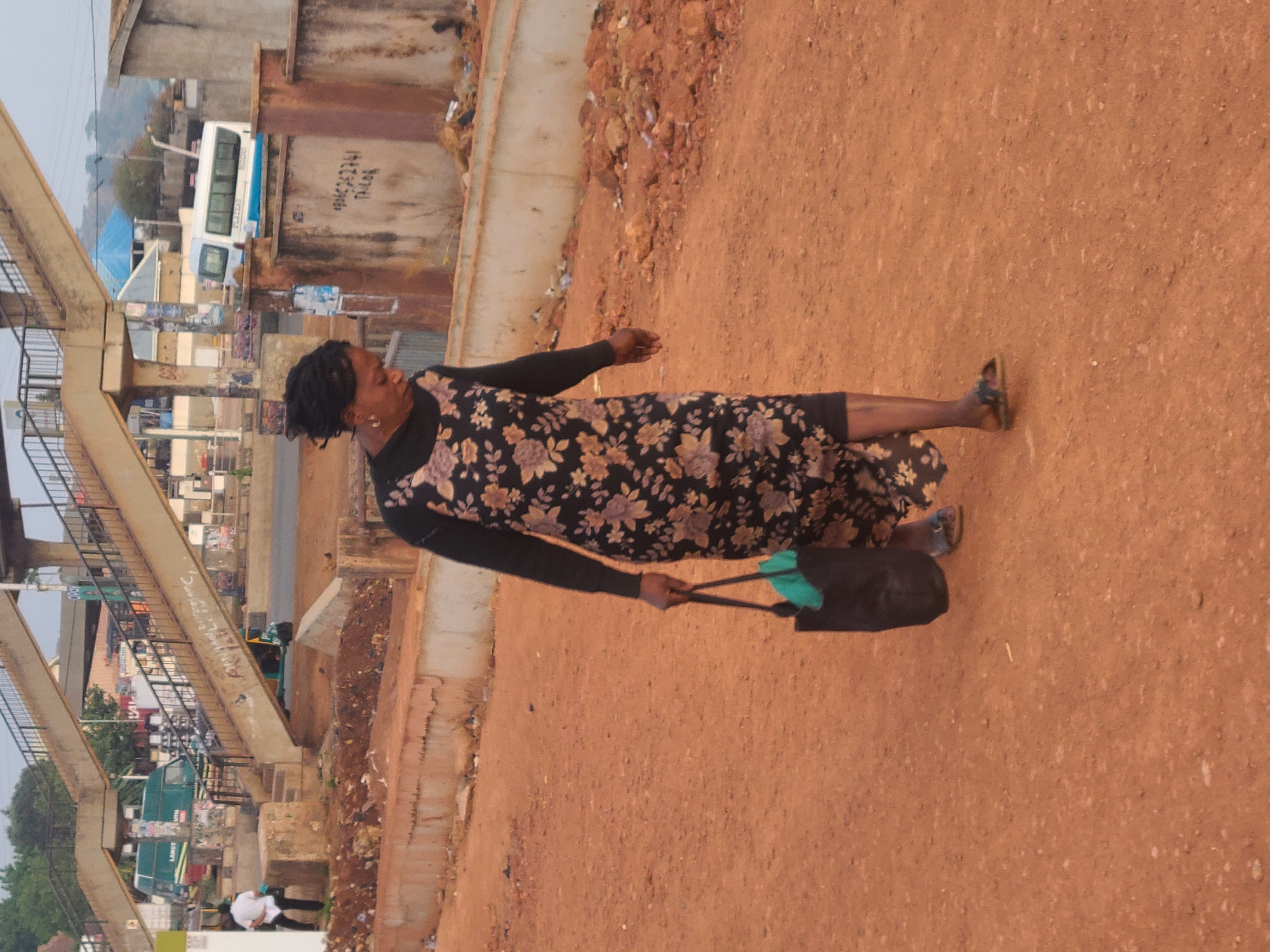 An adult woman is walking along a dirt road in Jos, Nigeria. She is carrying a black bag with what appears to be a pair of bright green socks. The woman is wearing a floral print dress and black long-sleeved shirt. The environment includes a pedestrian footbridge. A bus and other vehicles are visible in the background.