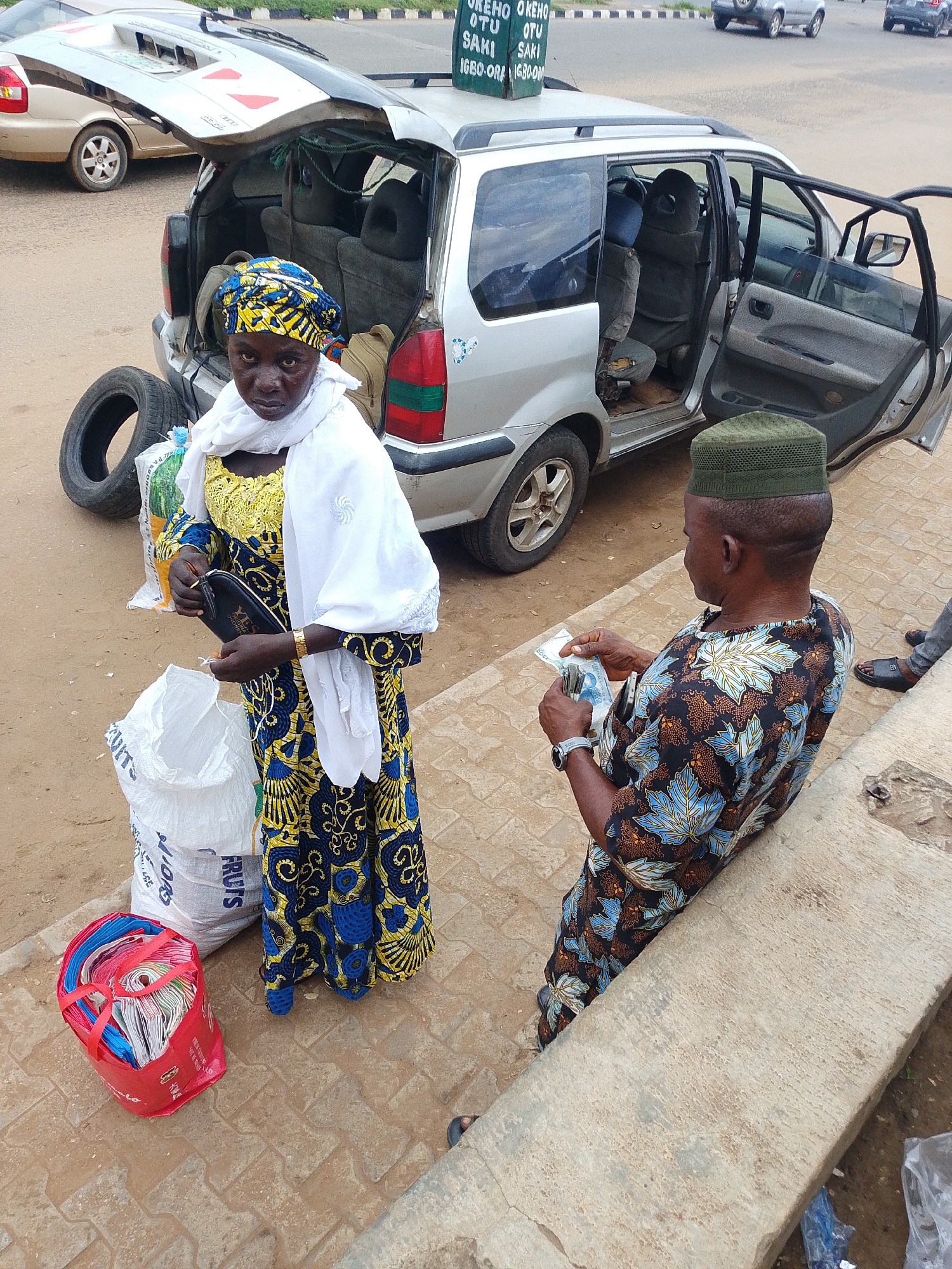 This media file captures a scene likely at a marketplace or roadside vending area in Nigeria, given the location context. The primary subjects are two individuals, a woman and a man, engaged in a transaction.
The woman, dressed in a vibrant blue and yellow patterned dress and a white headscarf, stands near the open back of a silver minivan. She is holding a black purse and appears to be either receiving or handing something to the man. Next to her are several large white bags, one with visible text that includes "FRUITS," and a red plastic bag filled with what appear to be folded shopping bags. A spare tire is also present near the minivan.
The man, wearing a green kufi cap and a patterned shirt, is seated on a concrete ledge. He is holding cash, suggesting he is making a purchase or receiving payment. His interaction with the woman is the central activity.
The minivan's rear door and one side door are open, indicating it might be used for transport or as a stall. The overall setting appears to be outdoors, with a paved surface beneath the subjects and a road with other vehicles in the background. A sign is visible on top of the minivan with some text, including "OKEHO," "OTU," and "SAKI," which are likely place names, possibly indicating the destination or origin of the goods or services being offered. The lighting suggests it is daytime. The scene is lively and functional, typical of commerce in a Nigerian setting.