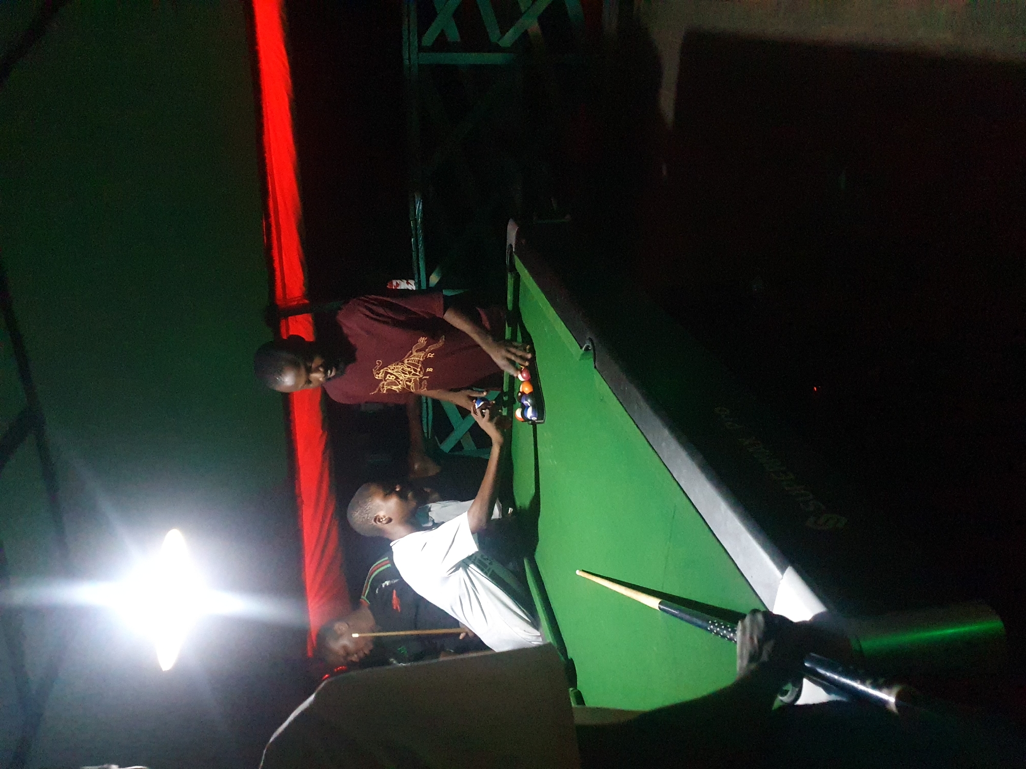 In Lokoja, Nigeria, a group of young men are gathered around a green pool table. The scene is dimly lit by a bright overhead light, casting long shadows. One man, wearing a burgundy t-shirt with a crest design, is poised to strike a billiard ball with a cue stick. Another man, in a white t-shirt, stands beside the table, looking towards the balls. A third man, in a black t-shirt, is partially visible behind him, also holding a cue stick. Several billiard balls are racked at one end of the table. The pool table itself appears to be a "Supermax Pro" model, as indicated by the lettering on its side. In the background, a red fabric is draped, adding a splash of color to the otherwise dark environment. The overall atmosphere suggests a casual game of billiards taking place at night.