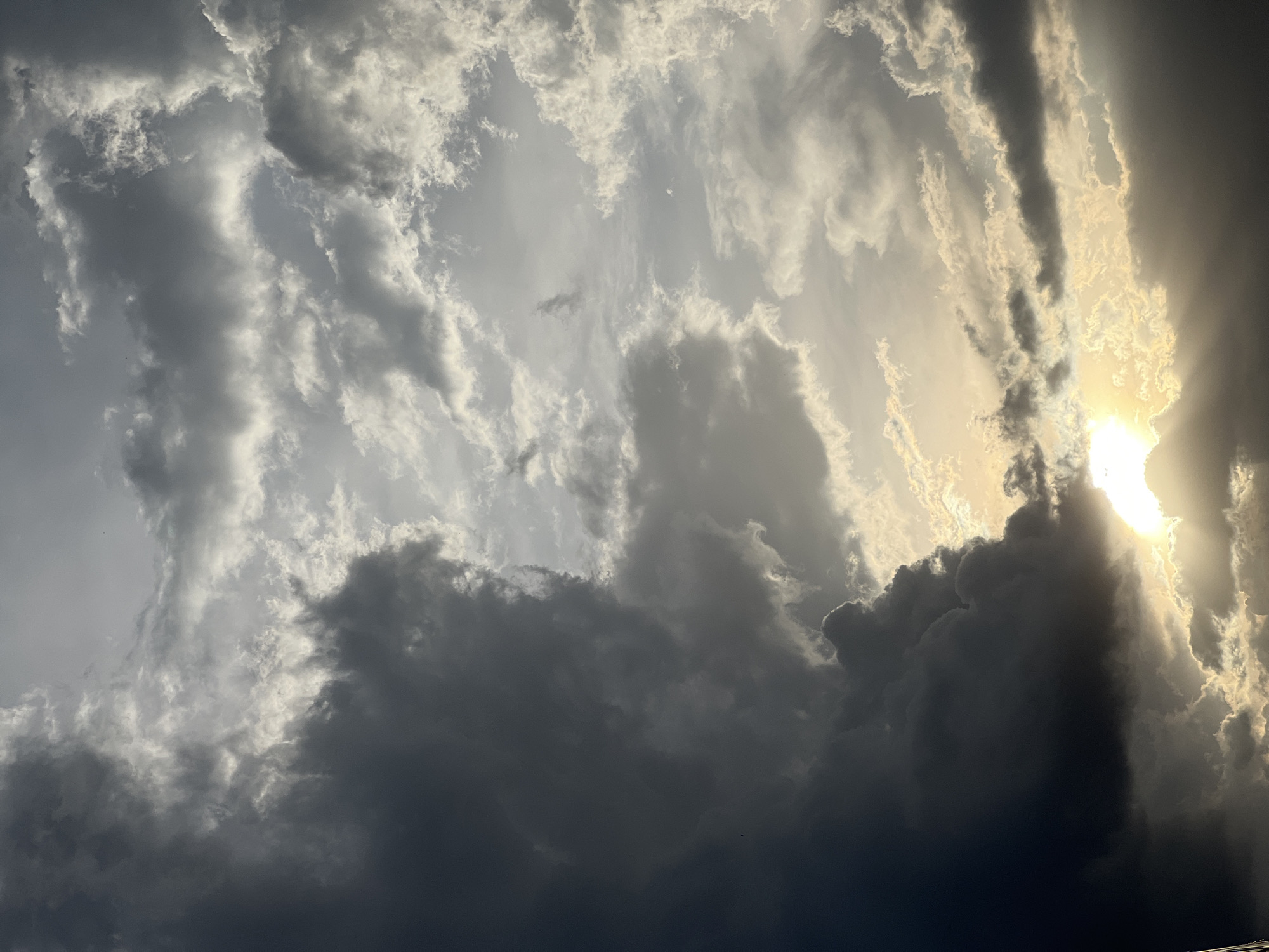 The image shows a view of the sky in Gura Top, Nigeria, with the sun partially obscured by heavy clouds. The clouds range in color from bright white to dark gray, with the sun appearing as a bright, diffused light source behind the clouds. The lower portion of the image is dominated by dark, ominous clouds, suggesting a potential storm. There are no people or objects other than the sky and clouds in the frame.