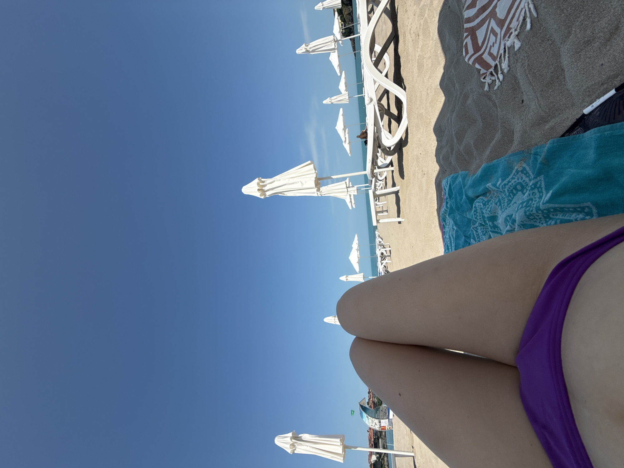 The image captures a first-person perspective of a sunny beach scene in Sozopol, Bulgaria. In the immediate foreground, the legs of a person are visible, wearing a vibrant purple bikini bottom, suggesting the viewer is relaxing on the sand or a lounge chair. To the right, a blue patterned towel and a brown and white patterned towel are laid out on the light-colored sand, with a dark object positioned nearby.
The scene extends towards the horizon with multiple rows of white beach umbrellas and matching white lounge chairs sparsely populated across the sandy shore. The clear, deep blue sky dominates the upper portion of the image, indicating bright, sunny weather. In the background, the calm blue sea meets the horizon. Strong shadows cast by the umbrellas and chairs onto the sand suggest it is midday or early afternoon. The overall impression is one of leisure and a vacation setting. No discernible text is visible within the image.