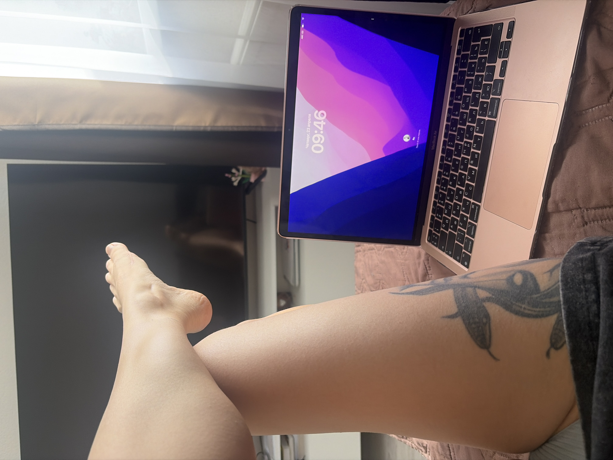 A first-person perspective image depicts a laptop computer open on a bed, next to a person's leg with a tattoo. The laptop screen displays the time "09:46" and the date, "Thursday, April 23," in Russian. The laptop is a pink or rose-gold color. The person's leg extends into the foreground, and a TV or monitor is visible in the background on the left side of the frame. The curtains are drawn closed, and a decorative object can be seen behind the laptop screen. The setting is indoors and appears to be a residential bedroom. The leg has a tattoo of a snake on the thigh. The bed is covered in a textured blanket.