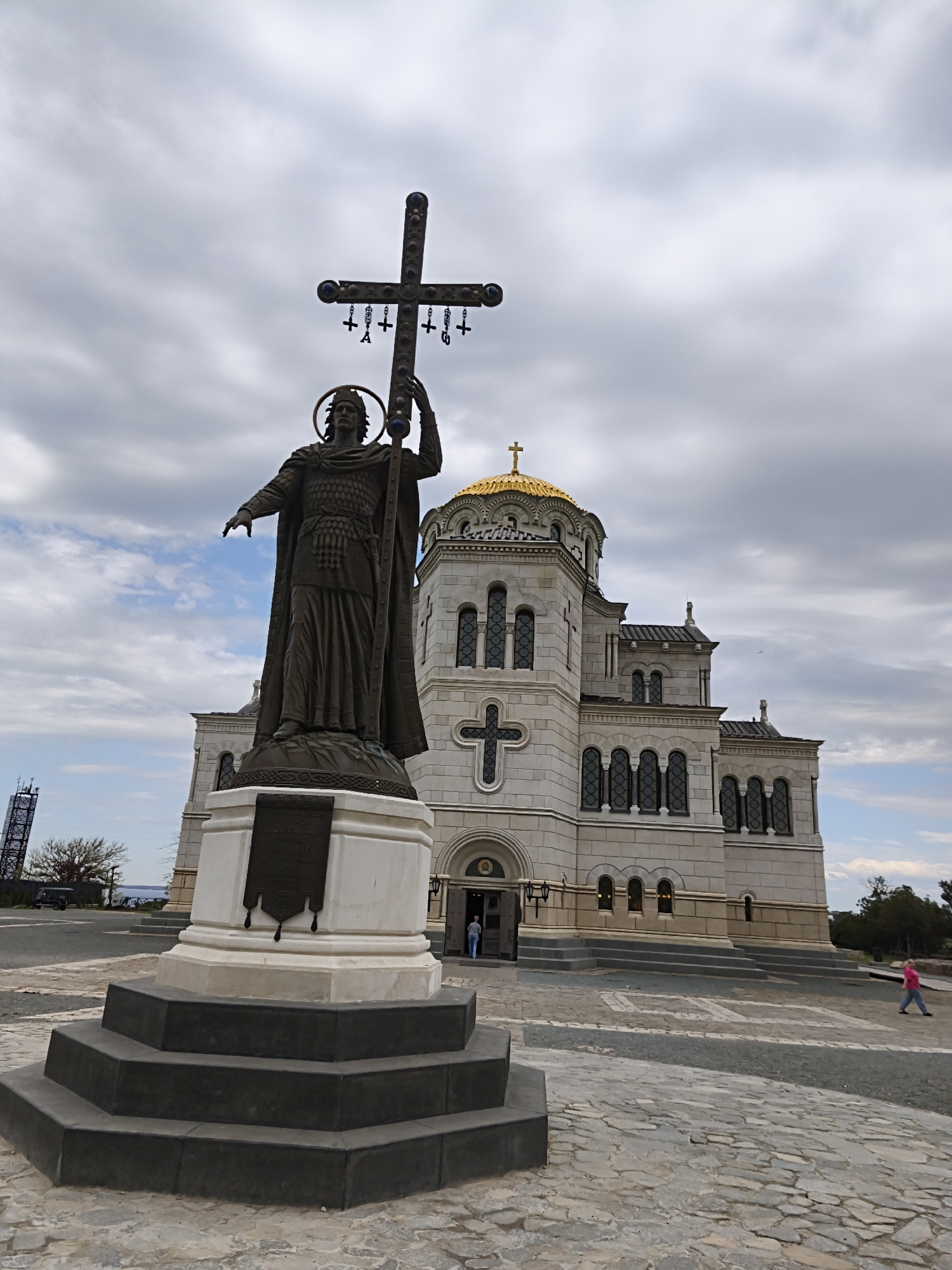 The image shows a statue of a man holding a cross, located in Sevastopol, Ukraine. The statue is bronze and stands on a multi-tiered stone base. The cross is adorned with smaller crosses. Behind the statue is a large, white stone building with a golden dome topped with a cross. The building has arched windows and a large cross on its facade. The sky is cloudy with patches of blue. A woman in a pink shirt is walking to the right of the statue.